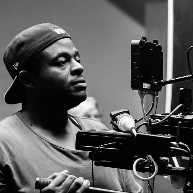 A man wearing a baseball cap, operating a film camera on a tripod, in a studio setting.