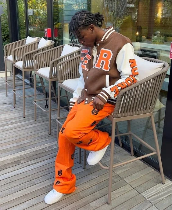 A young man sitting on a barstool at an outdoor seating area, wearing an orange and white varsity jacket with patches and lettering, orange pants with black logos, and white sneakers. He has short dreadlocks and is looking down.
