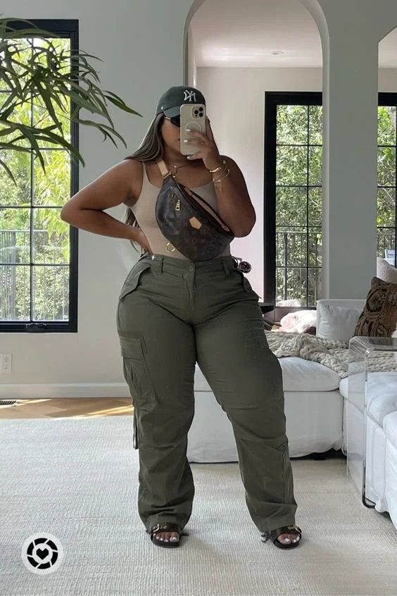 A woman taking a mirror selfie in a bright living room with large windows, wearing a black cap, sunglasses, beige top, olive cargo pants, sandals, and carrying a brown Louis Vuitton bag across her chest.