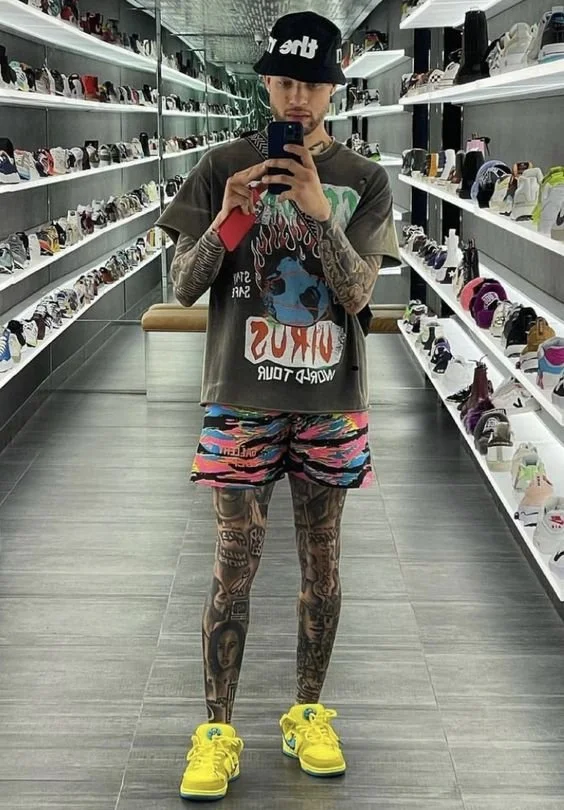Young man with tattoos is taking a selfie in a shoe store. He wears a black beanie, oversized t-shirt, colorful shorts, and bright yellow sneakers. Shoe shelves filled with sneakers line the walls behind him.