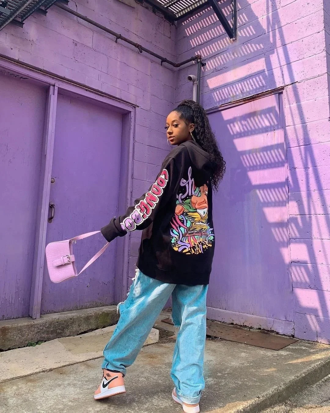 A young woman with curly hair in a high ponytail stands against a purple wall with purple door, wearing a black hoodie with colorful graphic art and text, ripped blue jeans, and white Nike sneakers. She holds a small pink handbag.