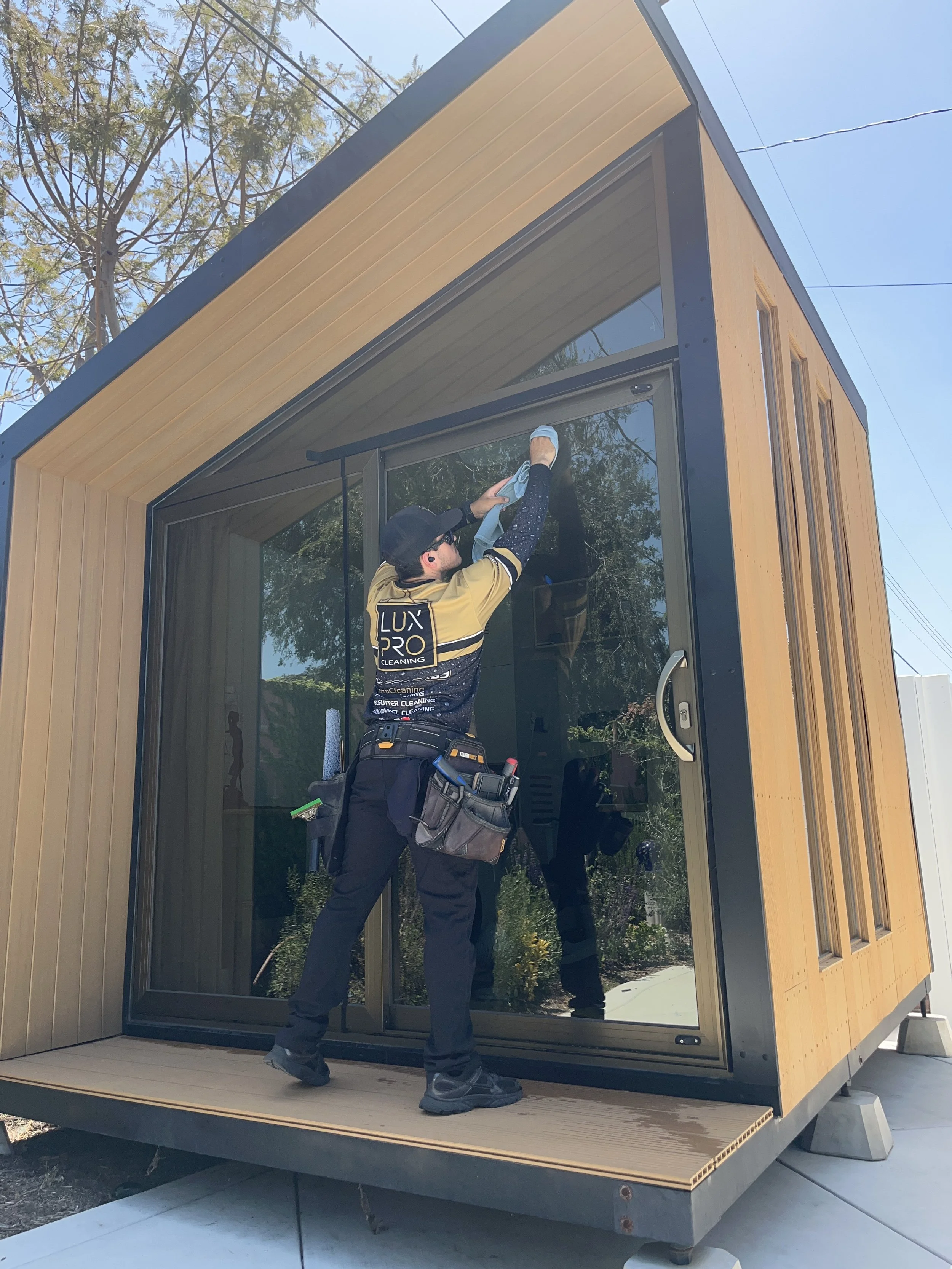 A worker cleaning large glass windows on a modern small building with wooden panel siding. The worker is wearing a yellow and black uniform with the logo 'LUX PRO CLEANING' and a black cap, and is using a cloth and spray bottle.