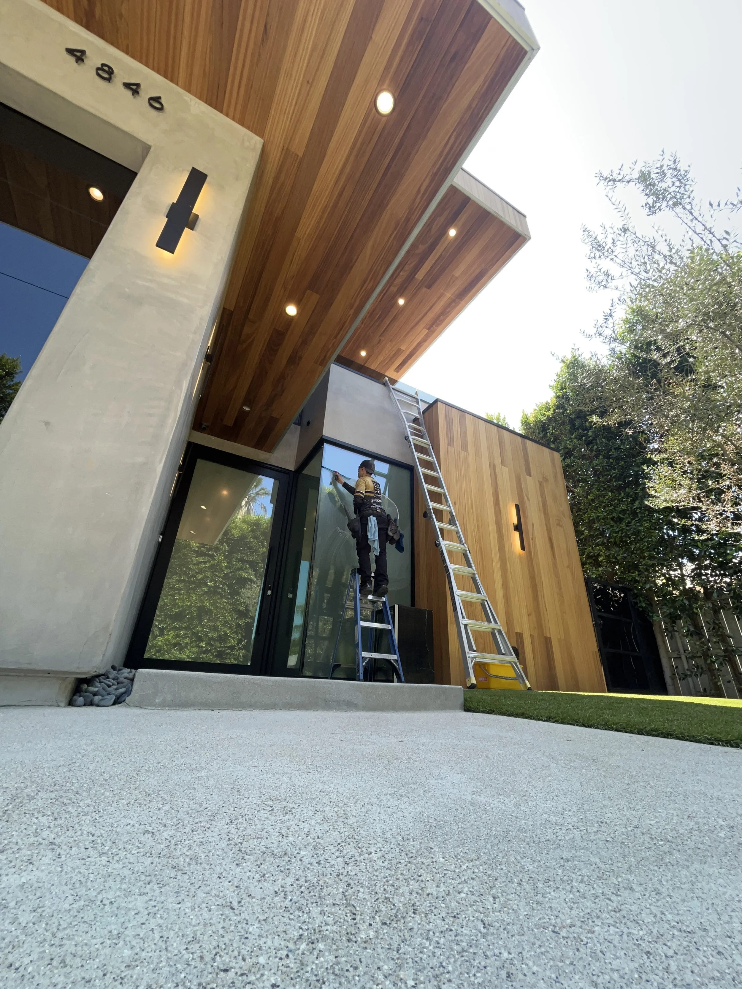 A person standing on a ladder cleaning the glass door of a modern house exterior with wooden and concrete elements, greenery, and a gravel pathway.