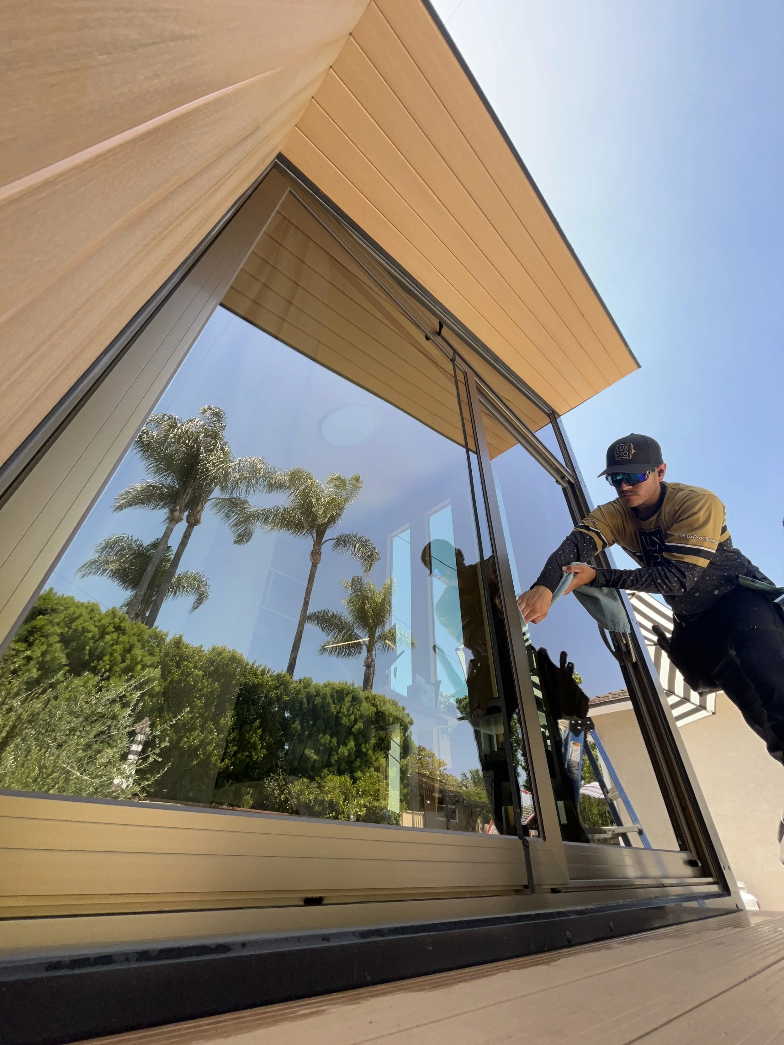 A worker is cleaning a large glass window on the side of a modern building with wooden panels and a roof overhang. The reflection of palm trees and the blue sky is visible in the window.