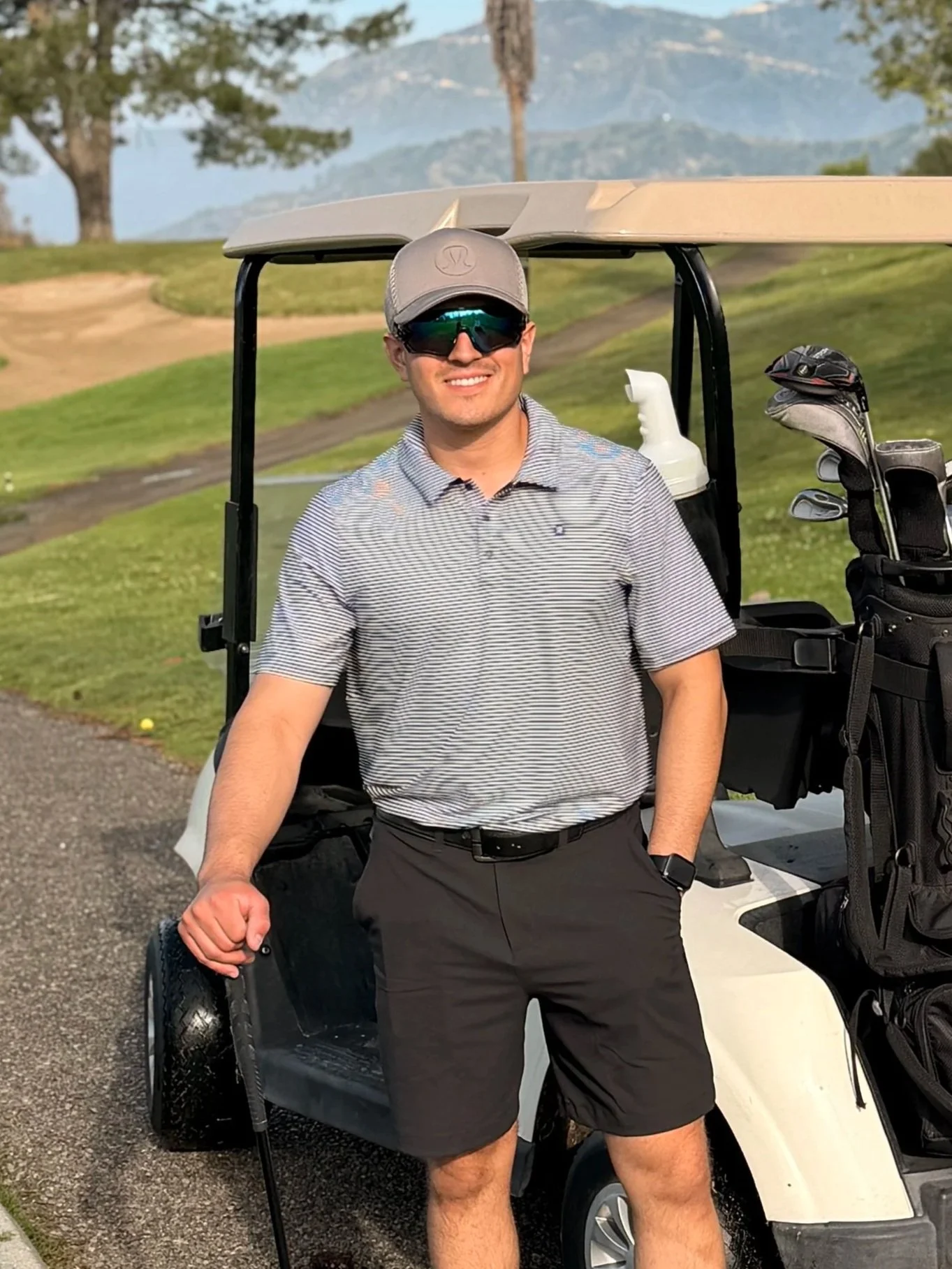 A man wearing sunglasses, a gray cap, a striped polo shirt, and black shorts standing next to a golf cart on a golf course with trees and mountains in the background.