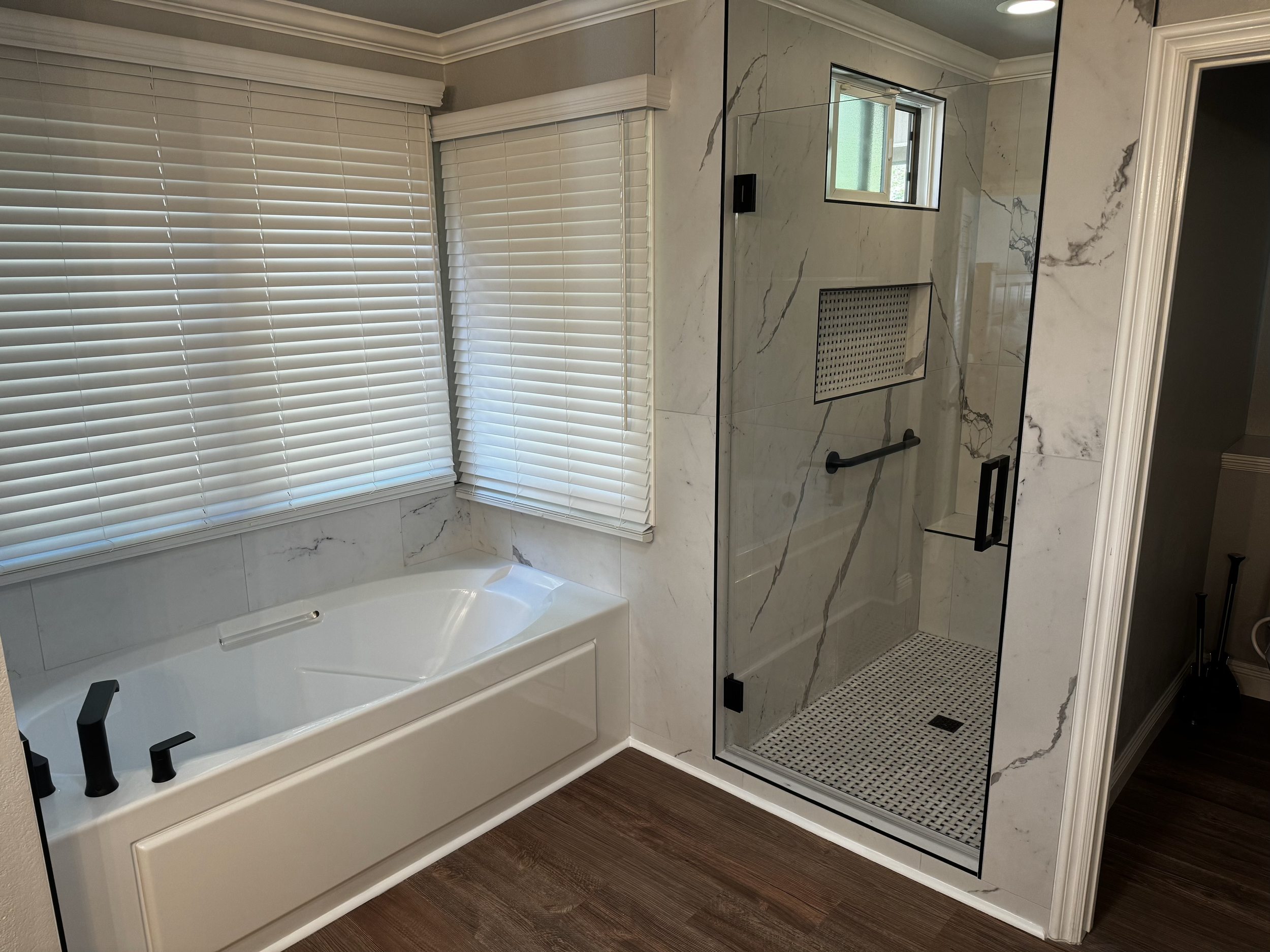 Modern bathroom with a soaking tub, large window with white blinds, marble shower with a glass door, and dark wood flooring.