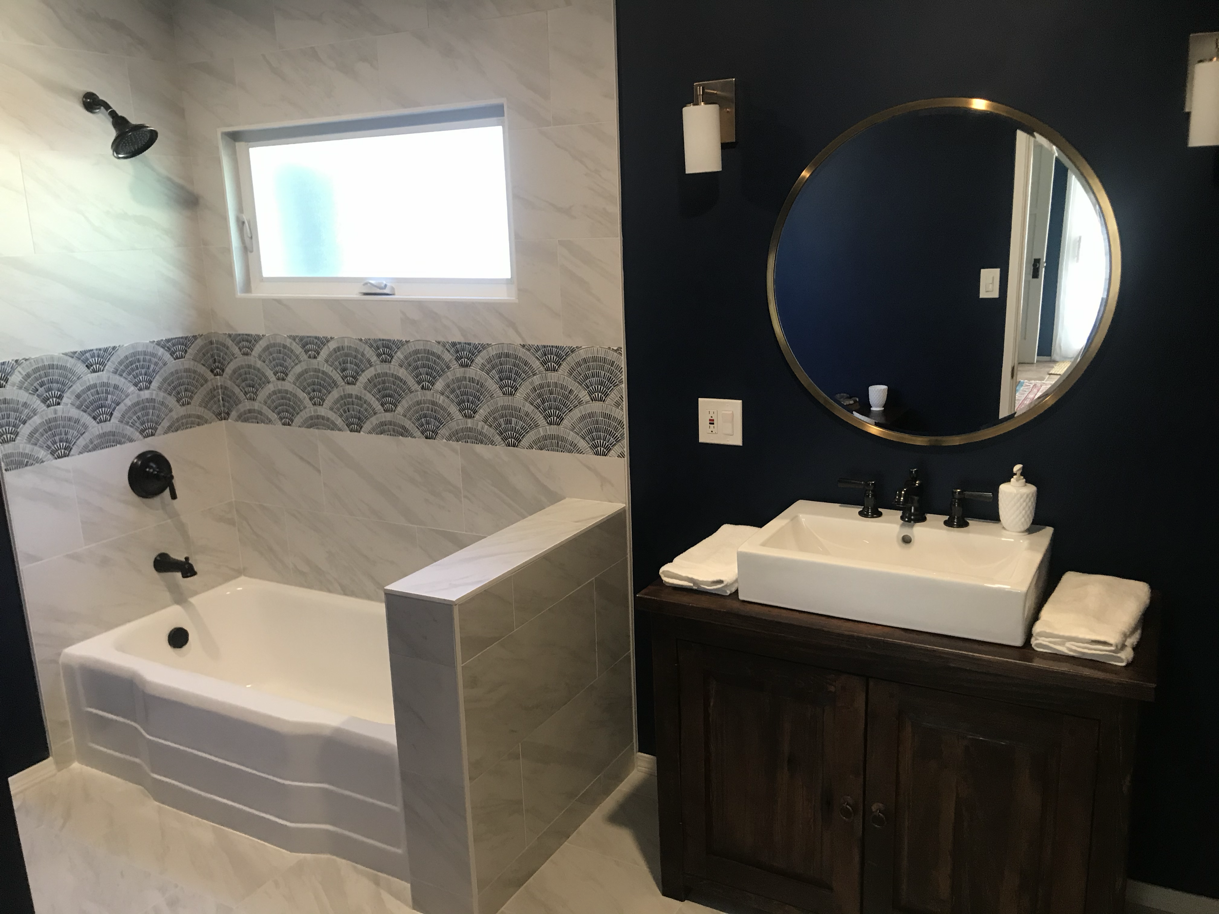 A bathroom with a walk-in shower with a black showerhead, a frosted window, a white bathtub, and a dark vanity with a white sink, black faucet, a mirror with a gold frame, and bathroom accessories on the counter.