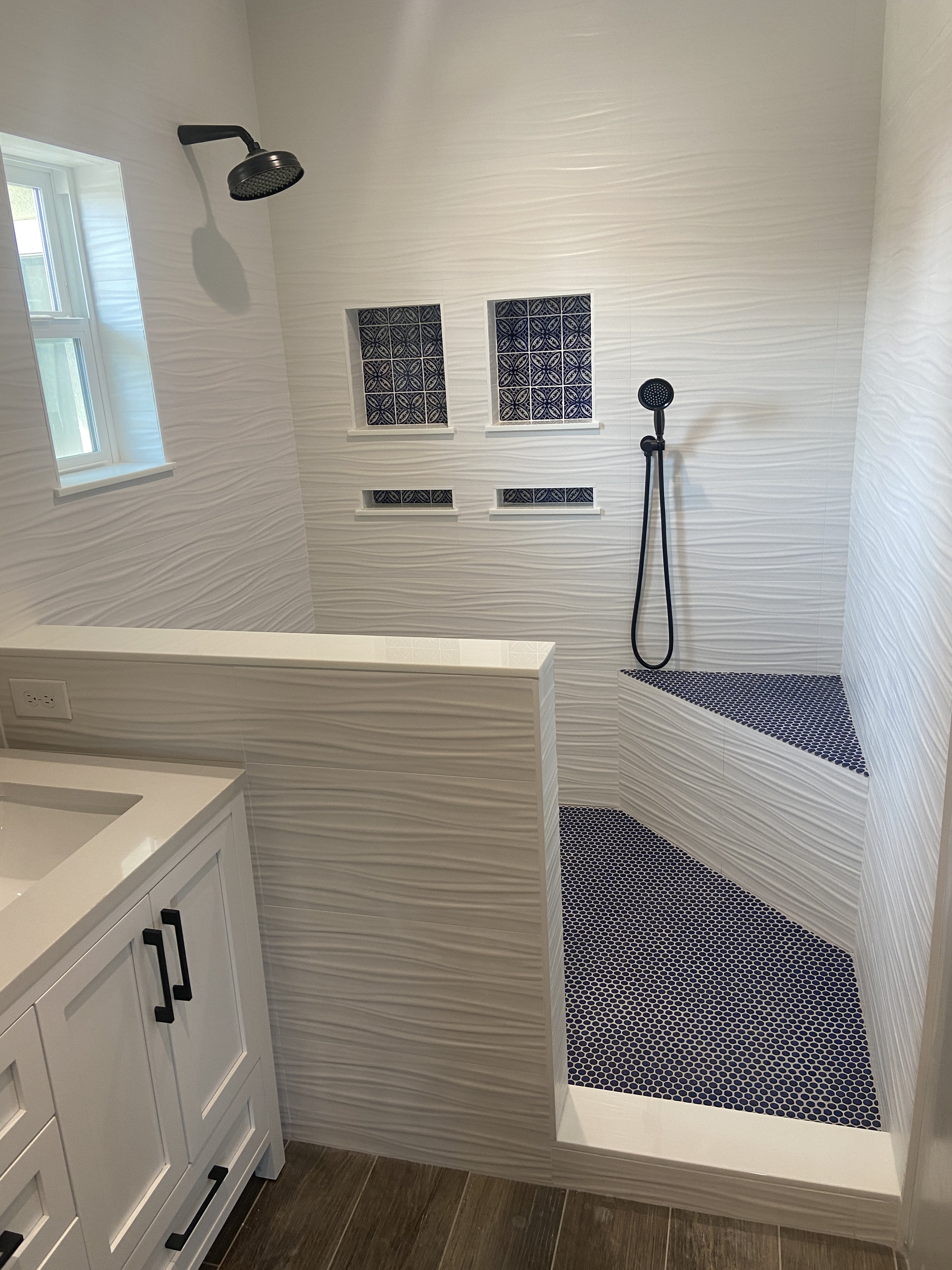 A bathroom with a walk-in shower featuring black and white patterned tiles, a black showerhead, handheld shower, a window, and a white vanity with black handles.
