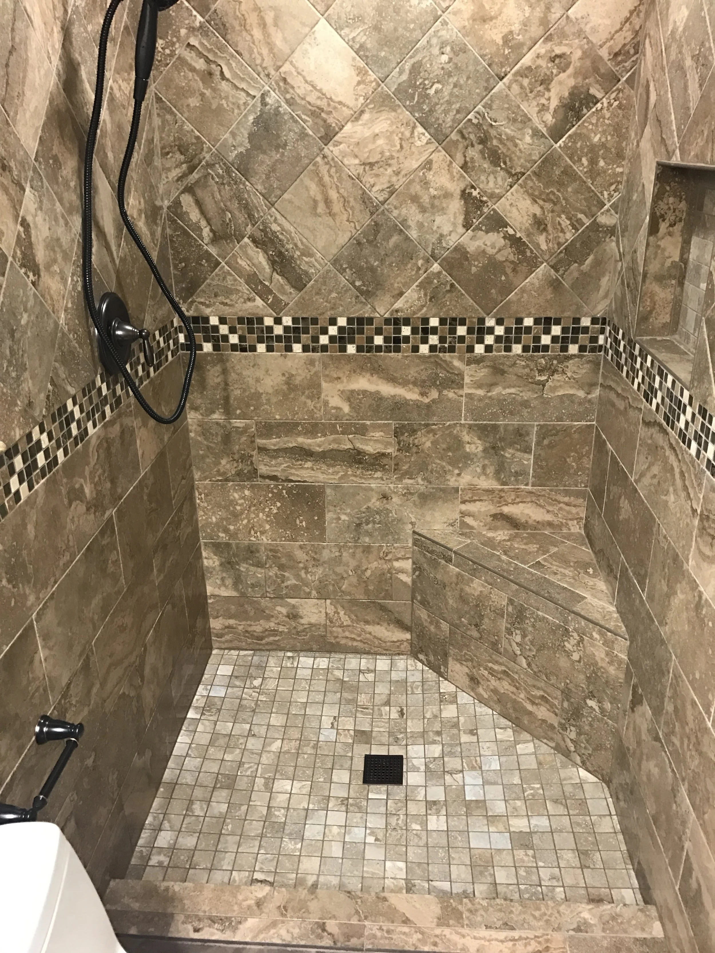 A tiled walk-in shower with a combination of large and small stone-like tiles, a black hand-held showerhead, a small built-in shelf, and a square drain on the floor.