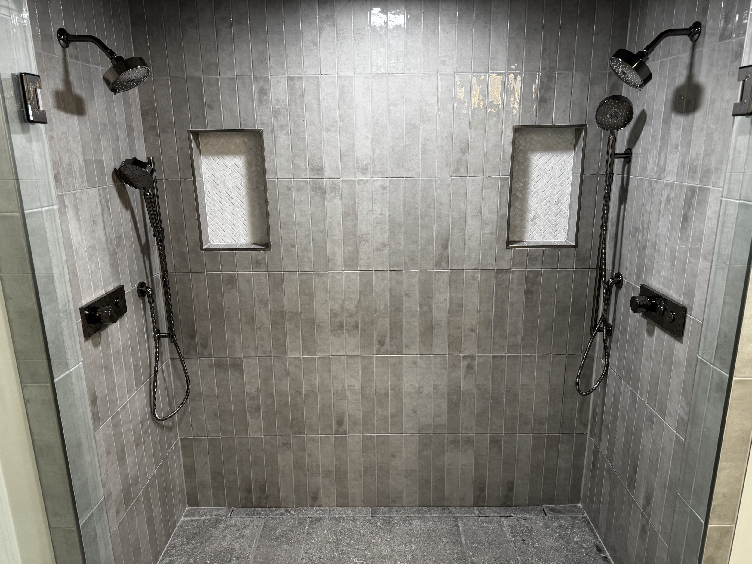 A shower area with two black showerheads mounted on a gray tiled wall, with two built-in wall niches lined with decorative textured tiles.