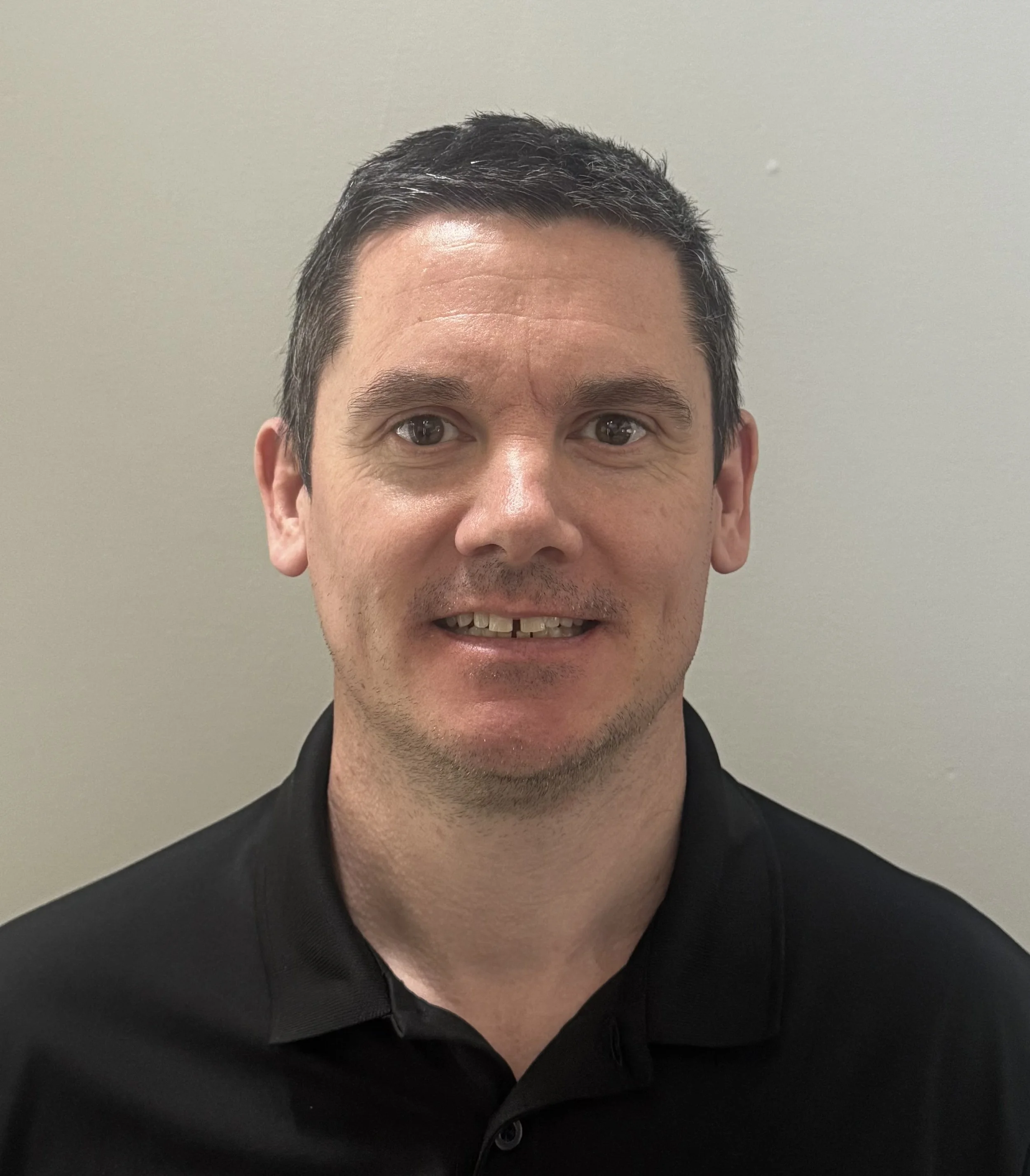 Close-up of a man with short black hair, brown eyes, and a light complexion, smiling slightly, wearing a black collared shirt, against a plain light grey background.