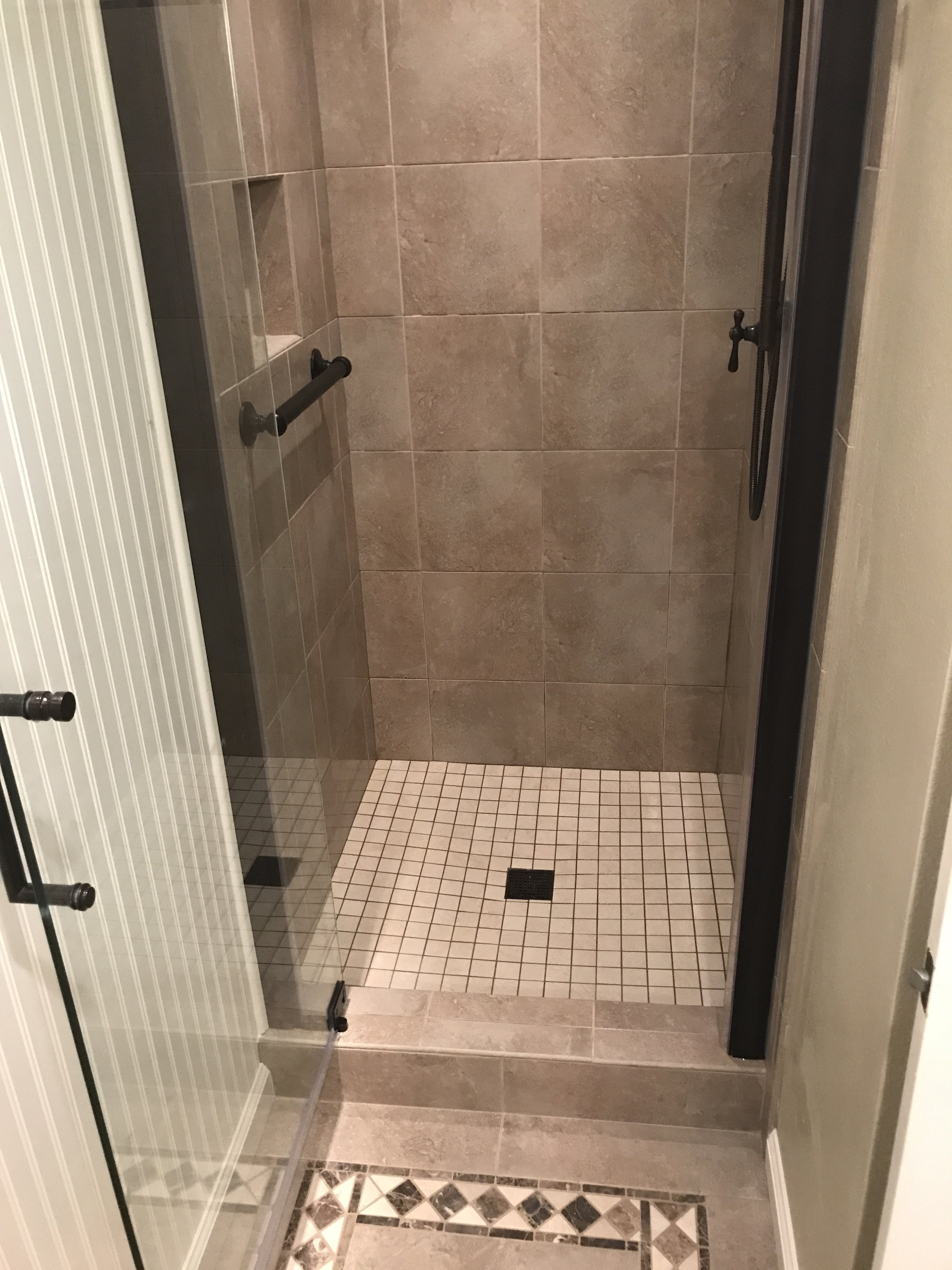 A bathroom shower with brown tile walls, a built-in shelf, a black grab bar on the left wall, a glass shower door with a black handle, and small square beige tiles on the shower floor leading to a black drain. The bathroom floor outside the shower ha