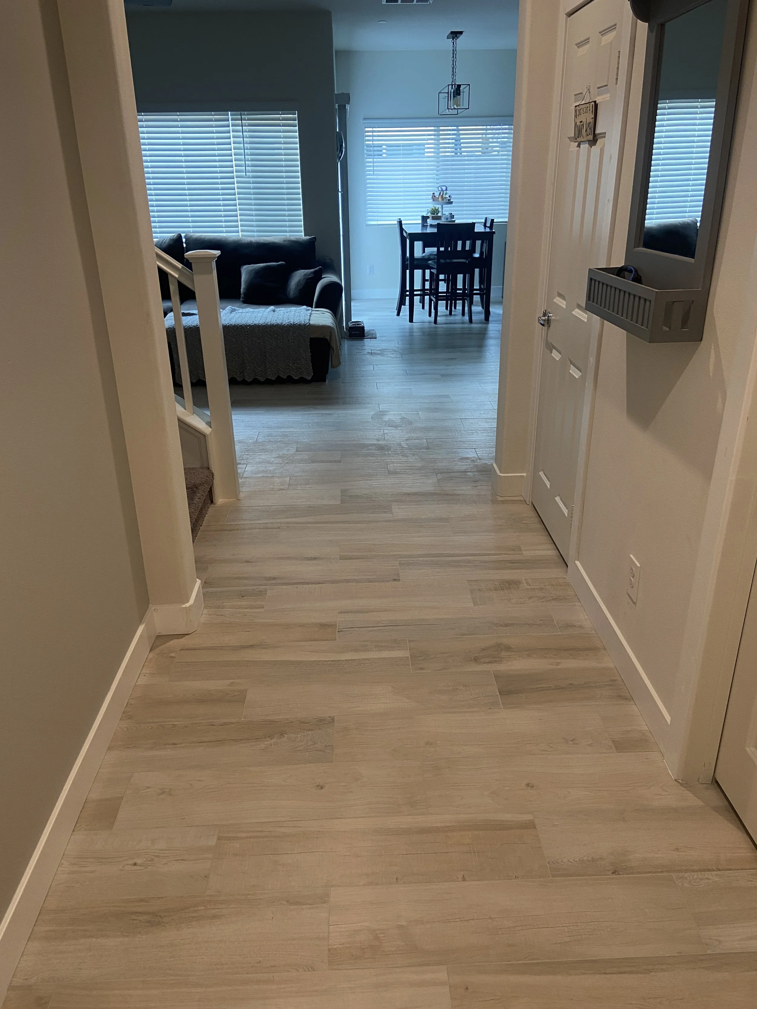View of a living room and dining area seen from the hallway. The floor is light wood, the walls are painted light cream. The living room has a window with closed blinds, a gray couch with pillows, and a blanket. The dining area has a rectangular tabl