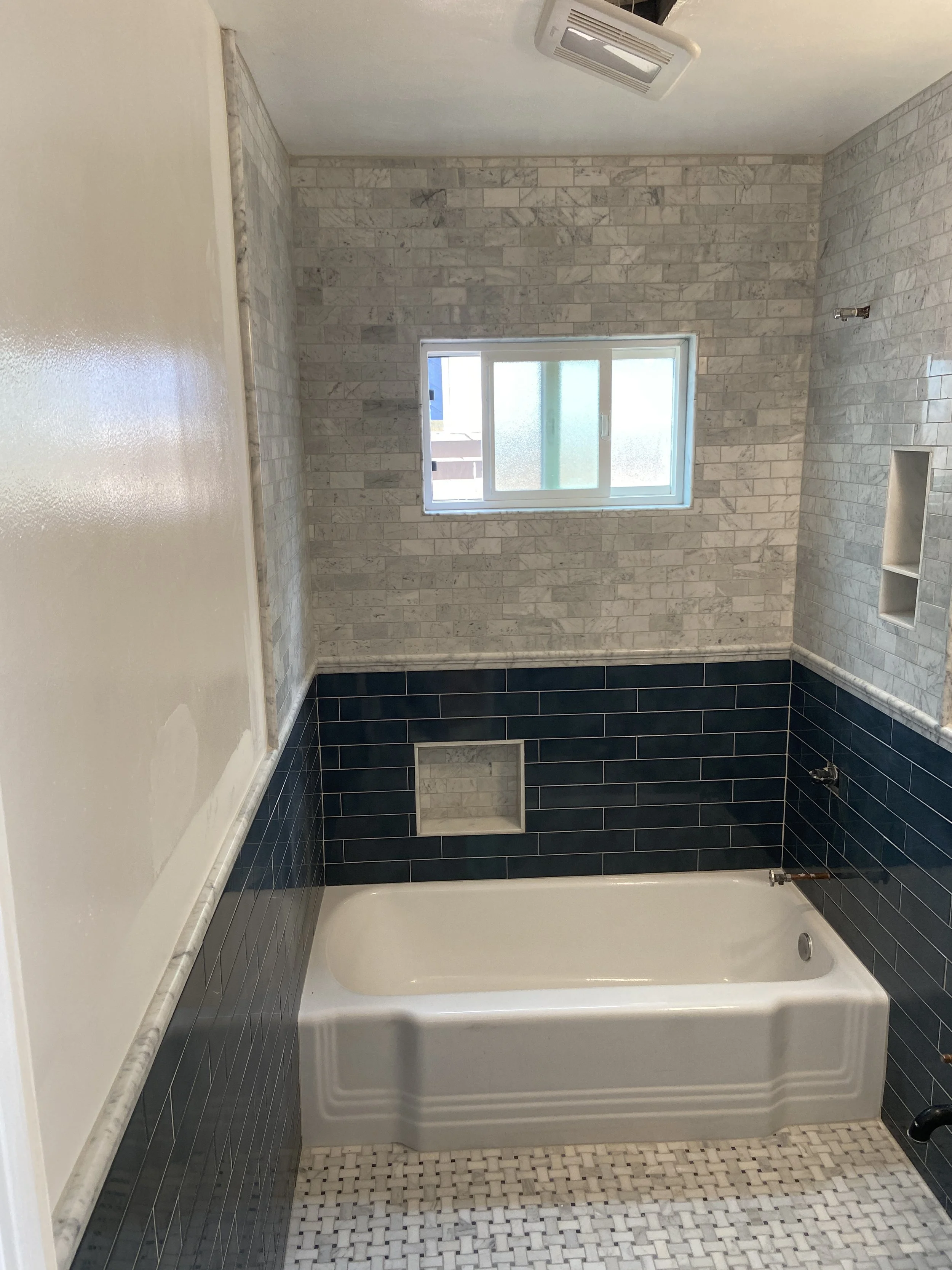 Bathroom with a white bathtub, navy blue tile wainscot, white and gray stone tiled wall, small square mosaic floor tiles, window, and built-in shelf.