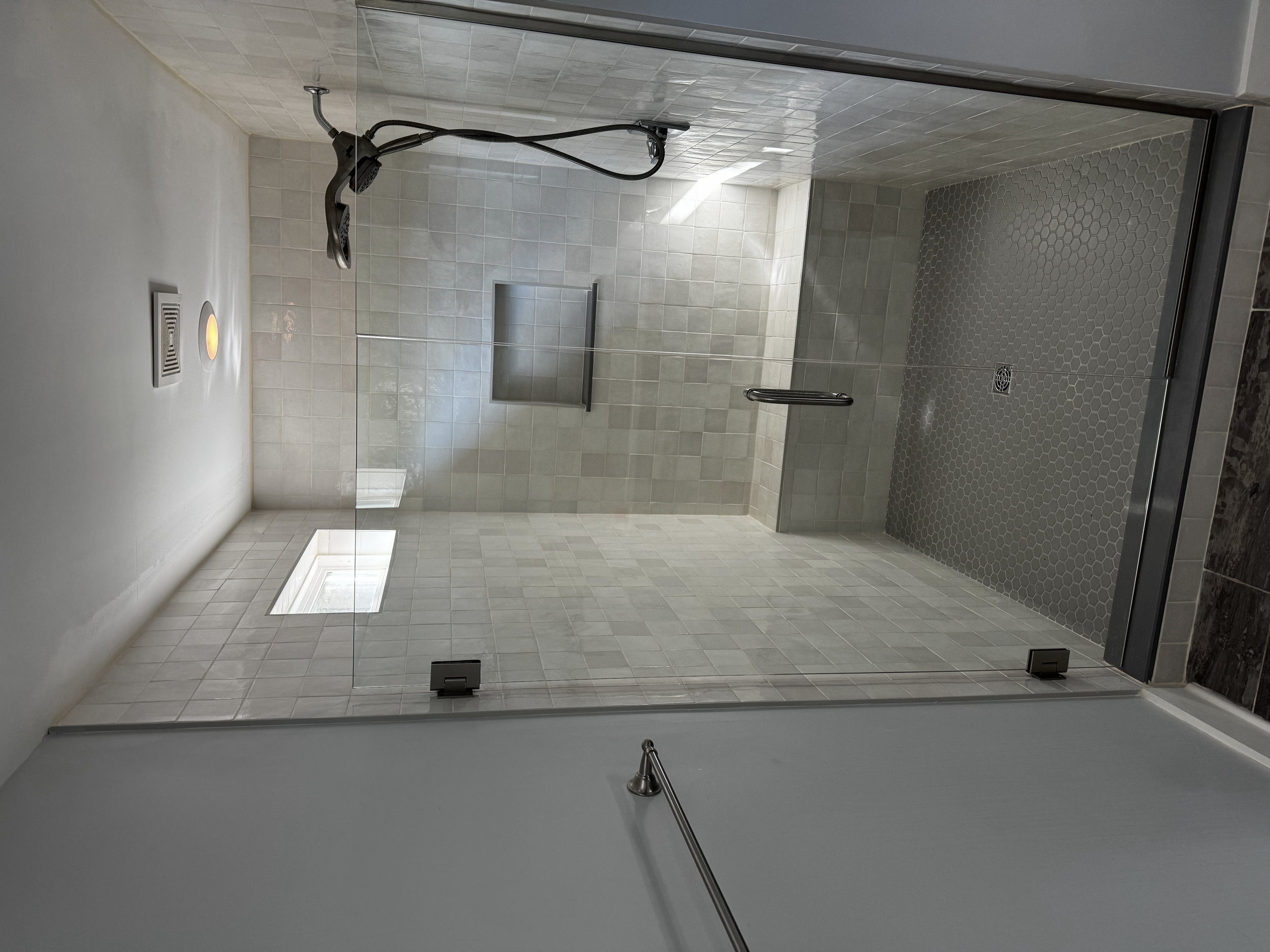 A modern walk-in shower with glass door, tiled walls, a handheld showerhead, a built-in niche for toiletries, a small window, and a floor drain.
