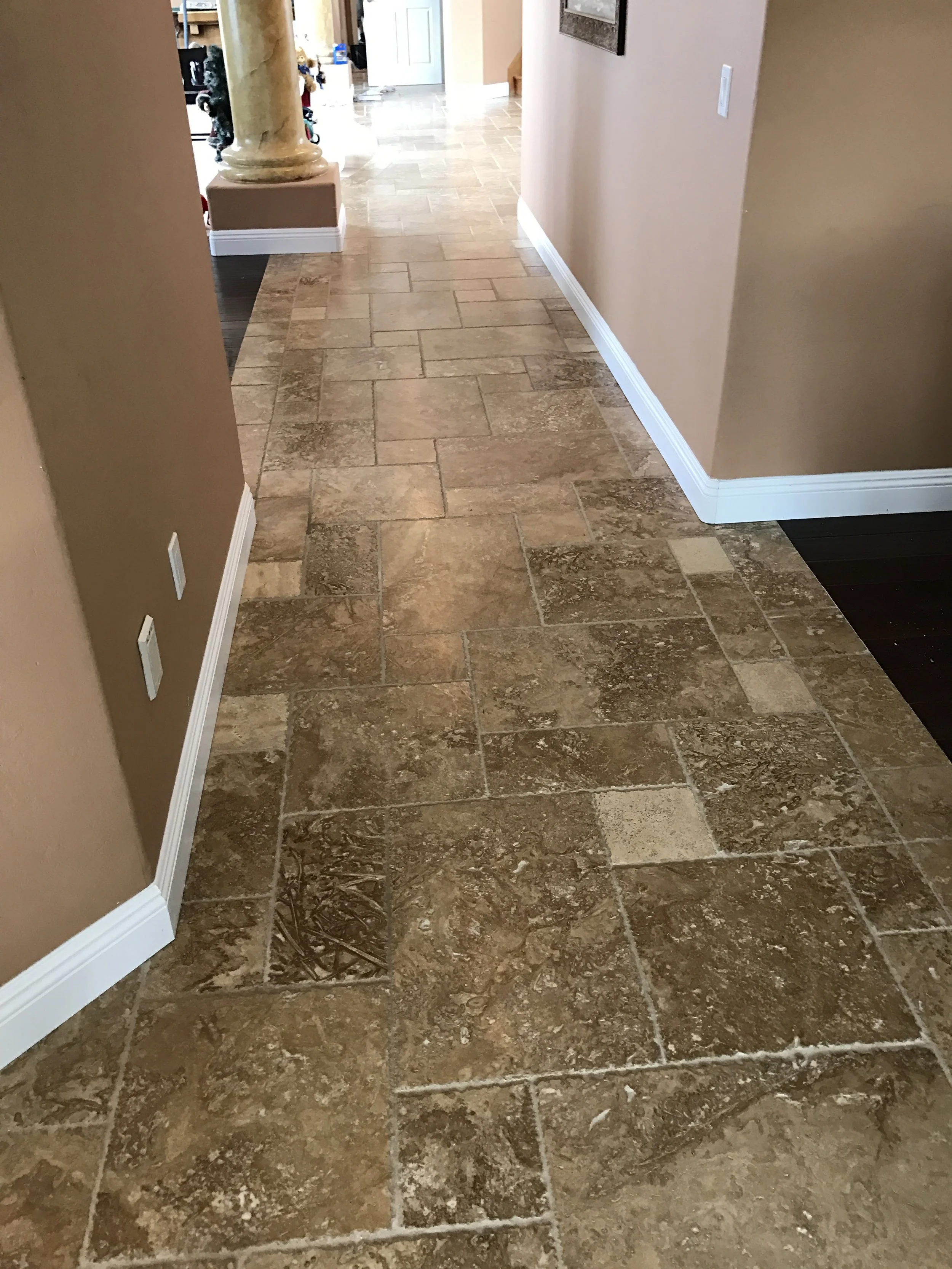 A tiled hallway with beige and brown tiles, white baseboards, beige walls, and a visible pole with a decorative base in the background.