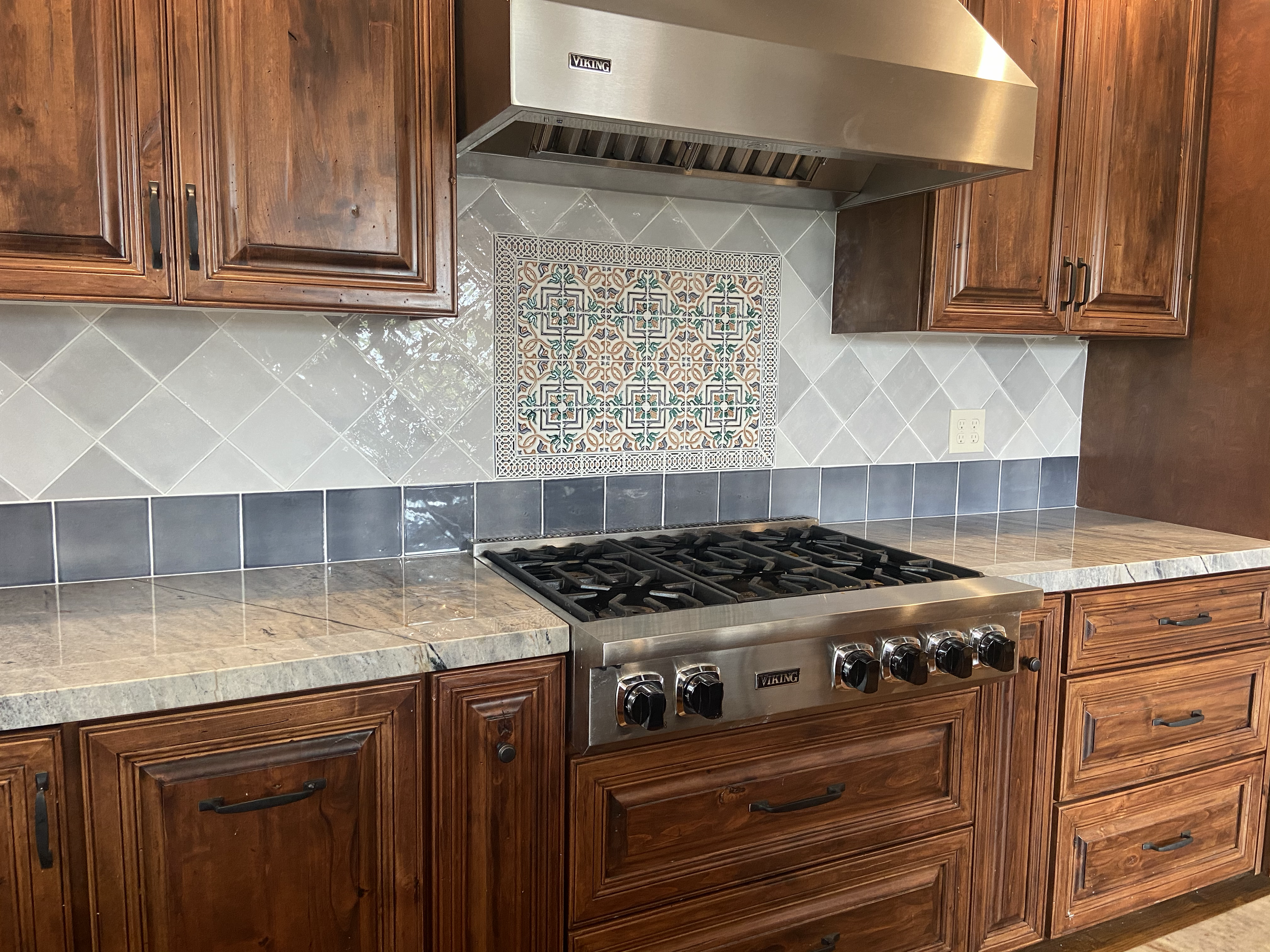 A kitchen with wooden cabinets, a stainless steel Viking vent hood, a patterned tile accent behind the stove, gray granite countertops, and a black stove with knobs.