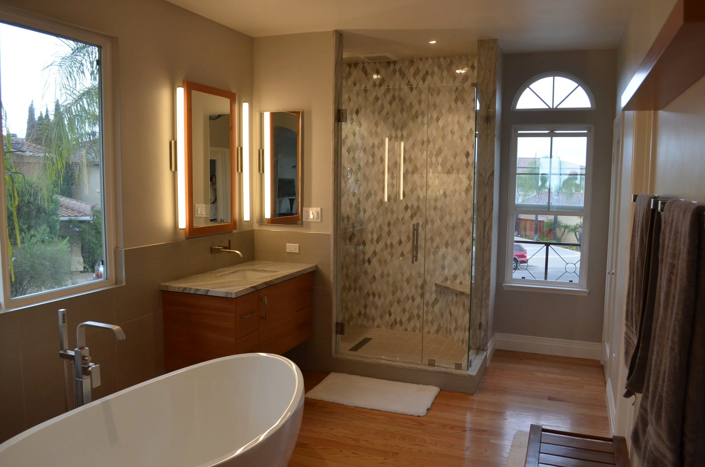 Modern bathroom with large windows, a freestanding bathtub, a walk-in shower with patterned tile walls, a wooden vanity with a marble top, two mirrors, and warm wooden flooring.