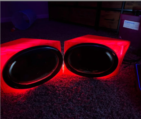 Two large red subwoofer speakers with black speaker cones, illuminated by red LED lights, placed on a carpeted floor.