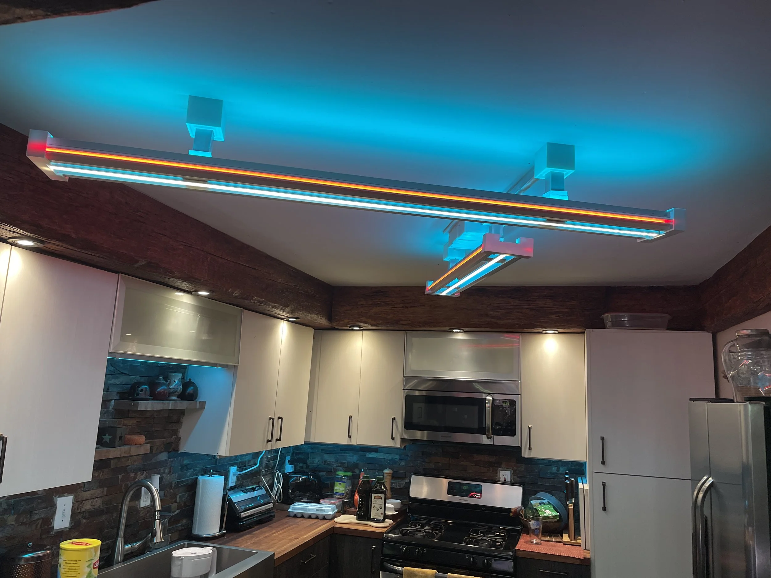 A modern kitchen with white cabinets, a wooden countertop, and a brick backsplash. The ceiling features colorful LED strip lights, and various kitchen appliances and items are visible, including a microwave, coffee maker, and utensils.
