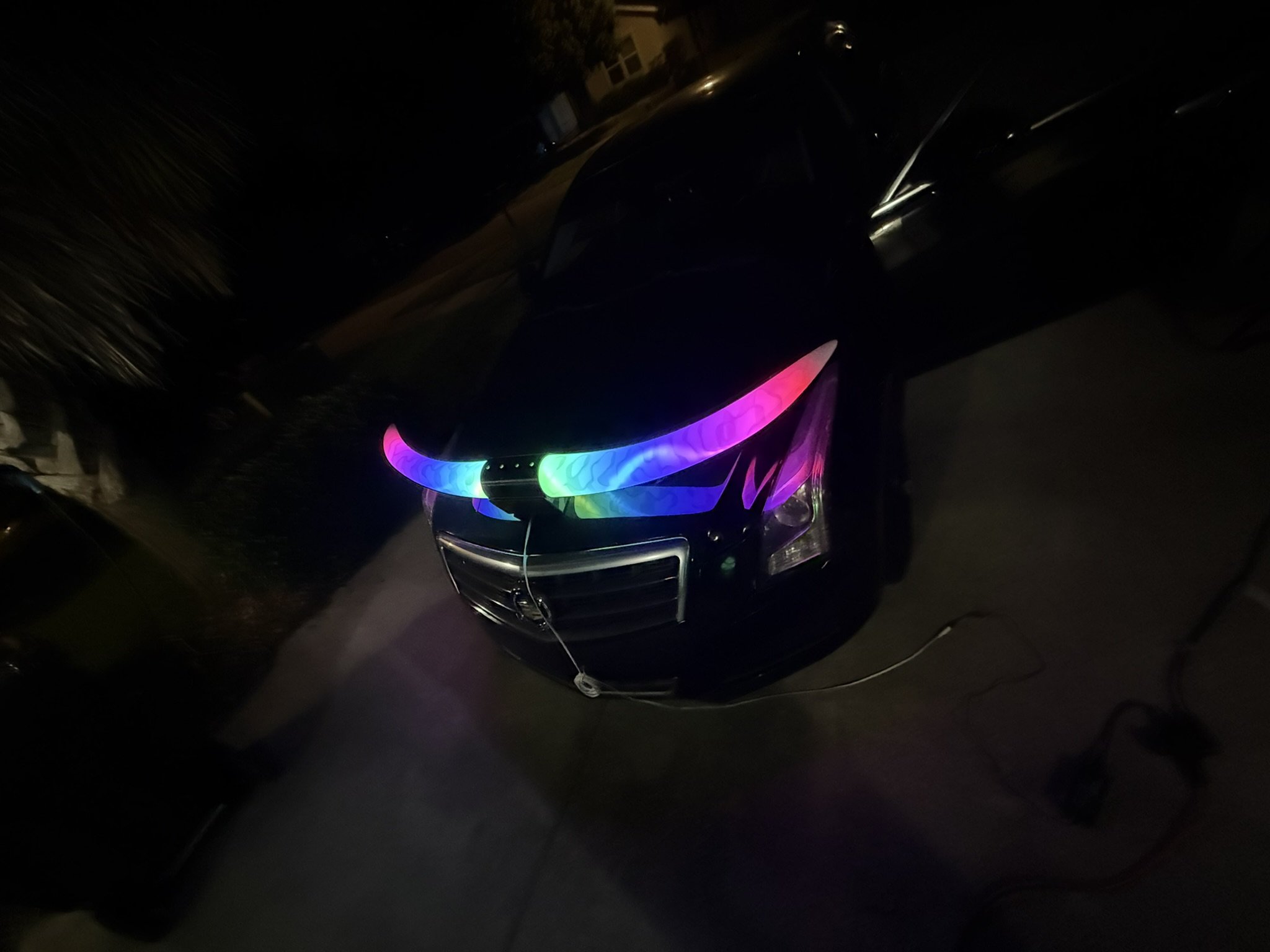 A black car with illuminated, rainbow-colored LED lights on the front grille parked outdoors at night.