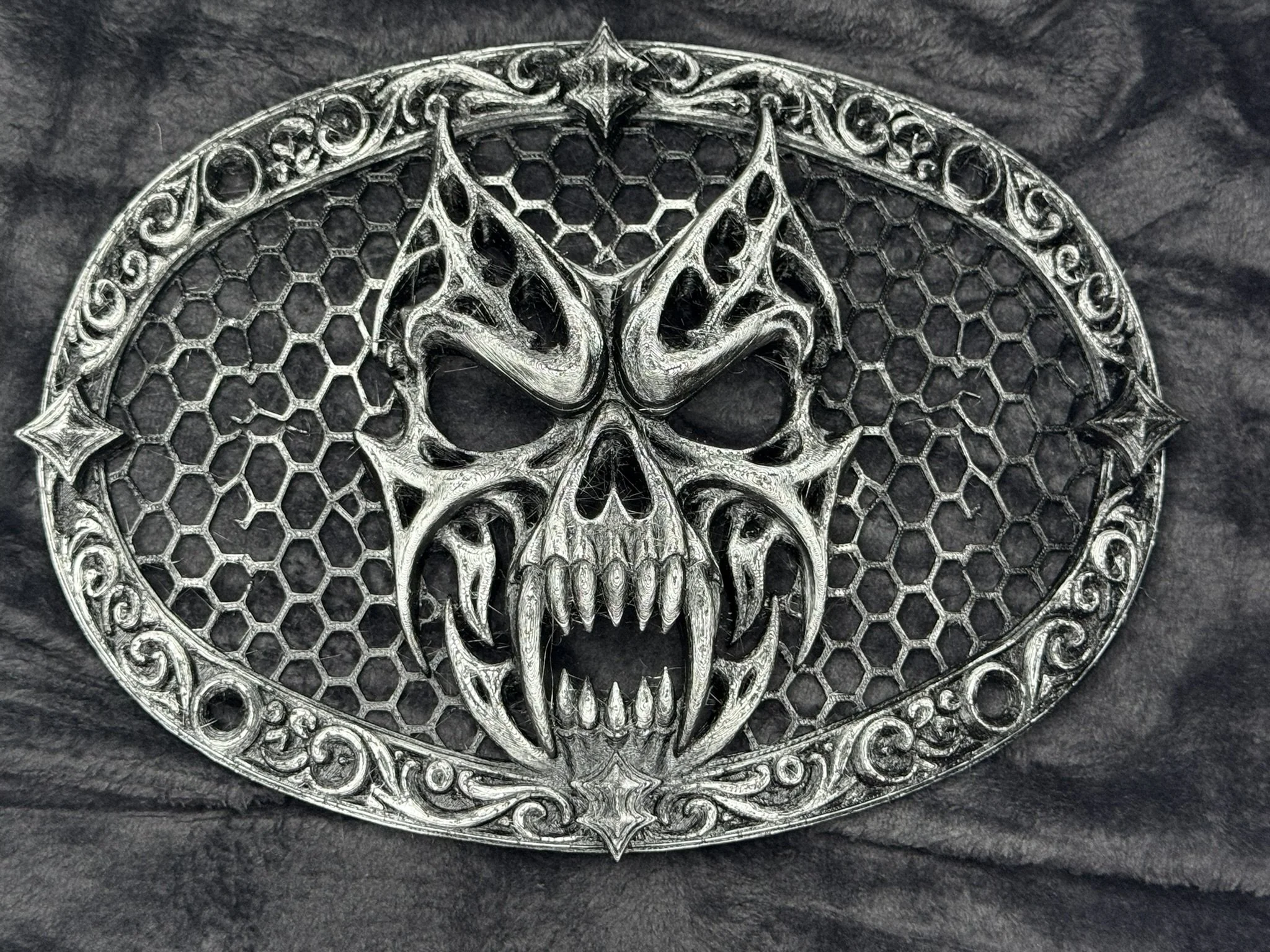 A metallic emblem featuring a skull with intricate design elements, surrounded by a honeycomb pattern and decorative borders.