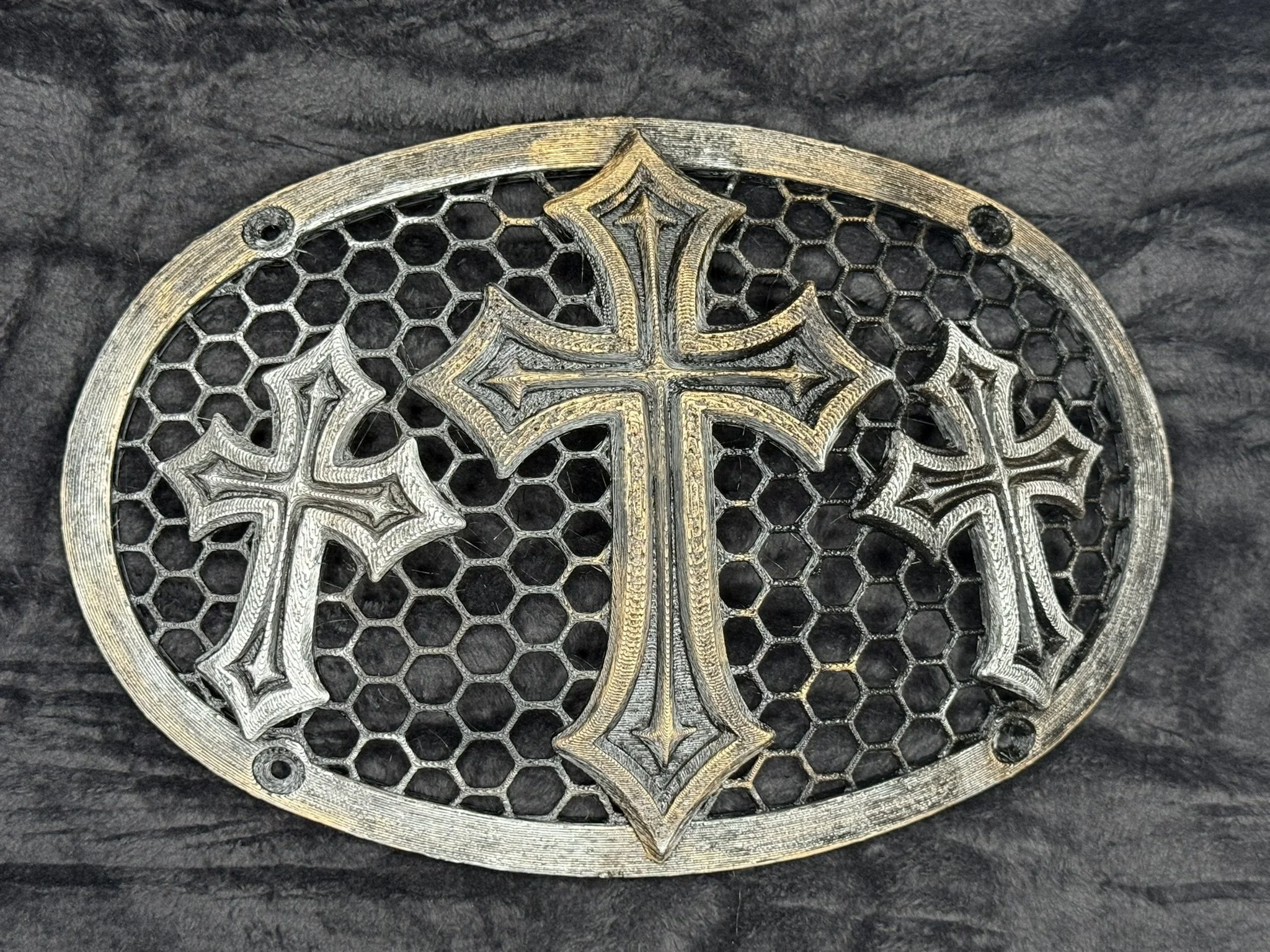 Close-up of intricately designed metallic emblem or jewelry piece with cross and decorative patterns, placed on a dark textured surface.