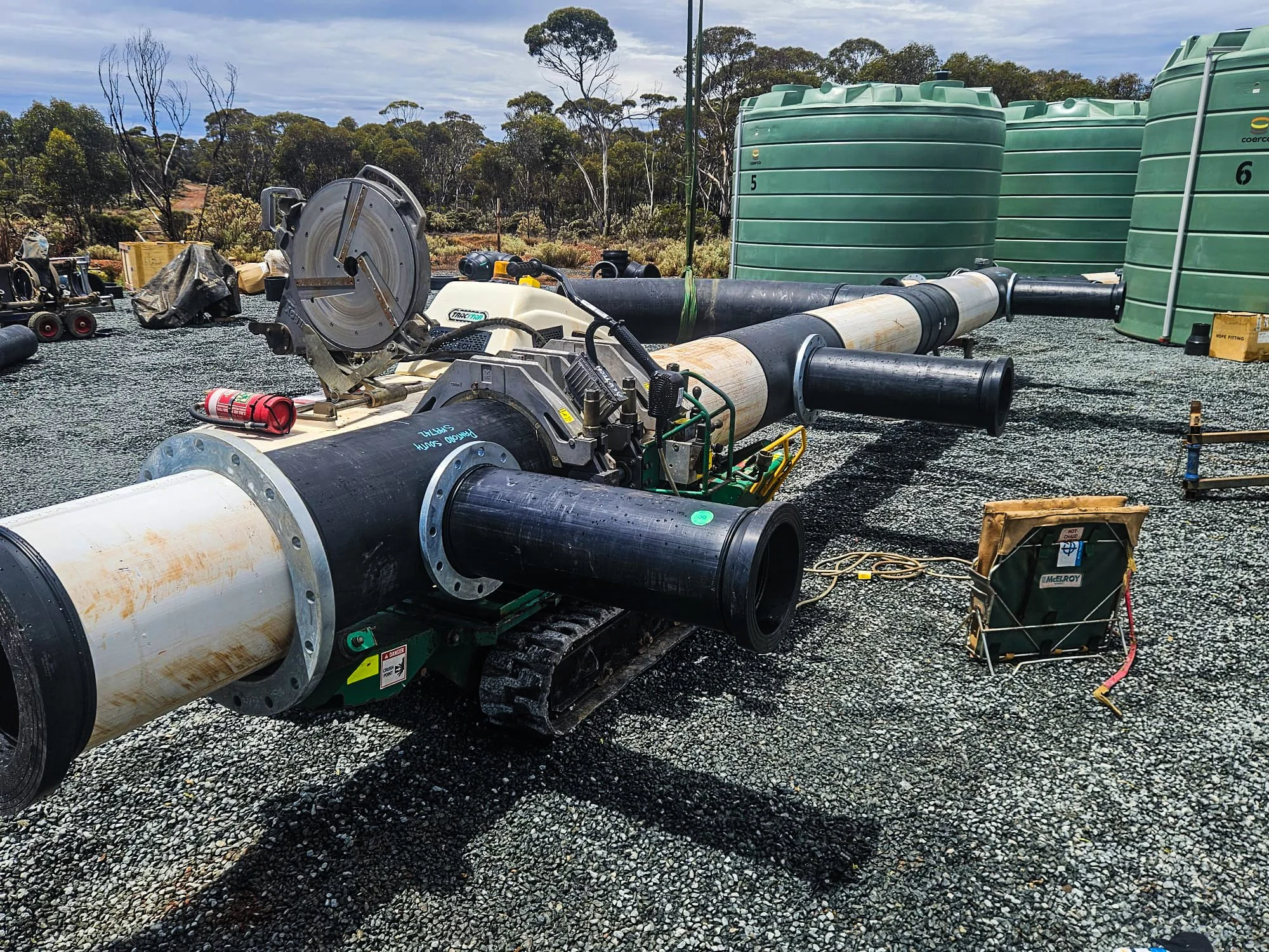 Construction site with large pipes, green water tanks, and equipment on a gravel surface under cloudy sky, surrounded by trees.