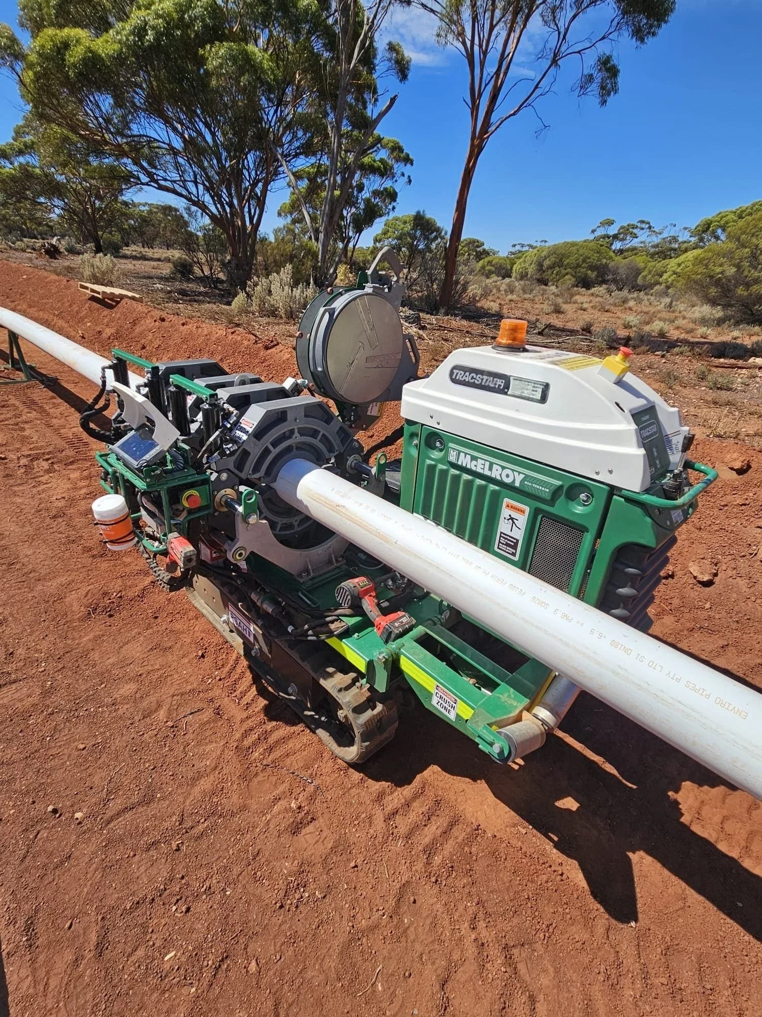 A pipeline construction machine on red dirt with trees and a blue sky in the background.