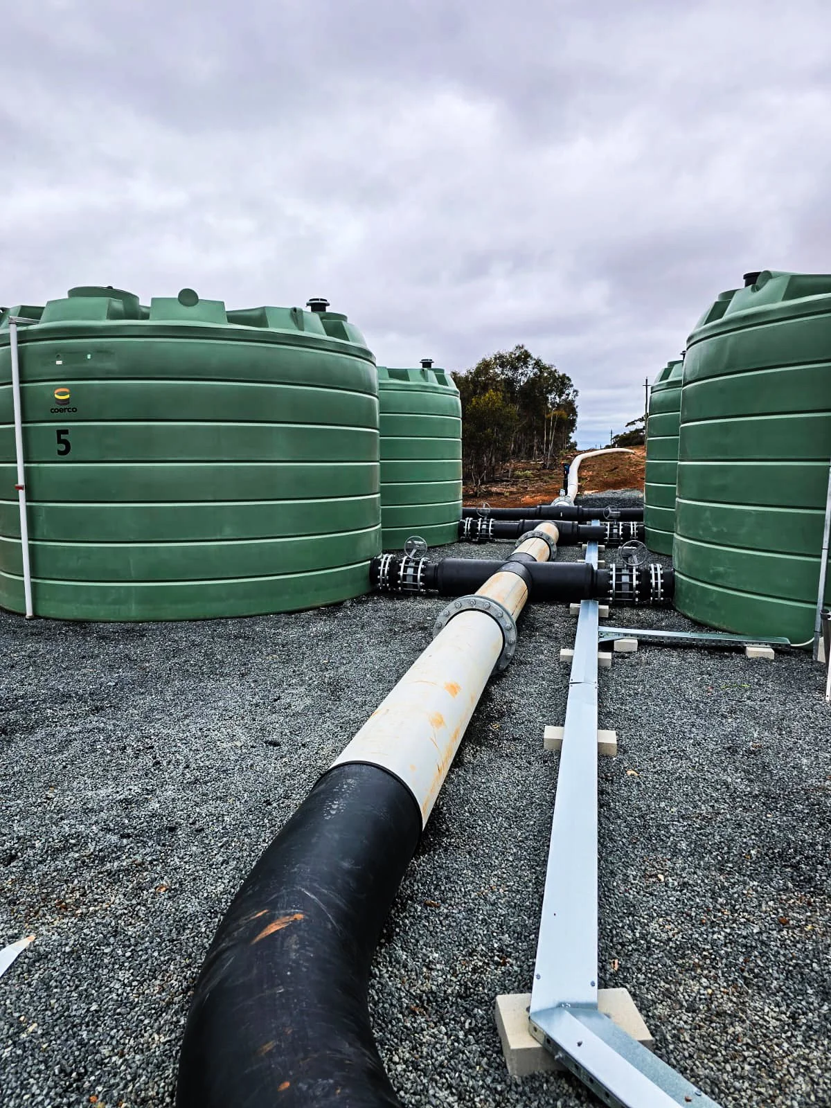 Large green water or chemical storage tanks with attached pipes and gravel ground, and a cloudy sky.