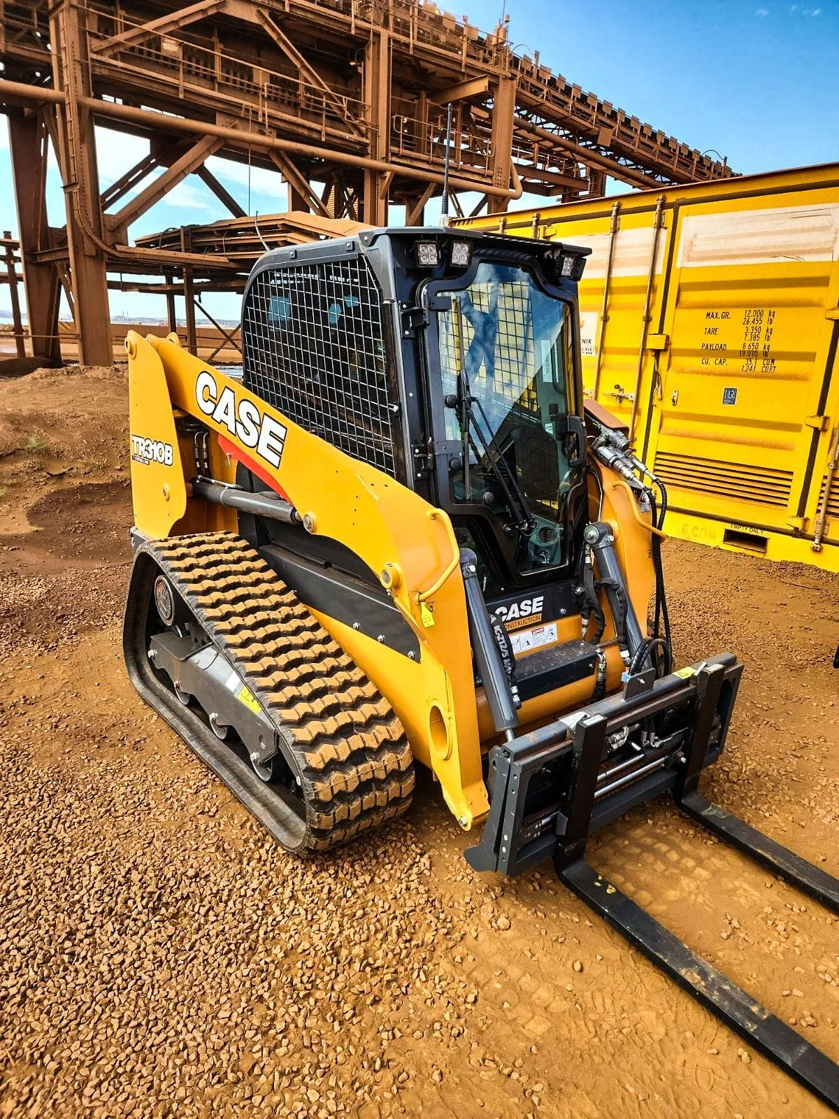 A yellow CASE compact track loader with black tracks parked on a dirt surface at an industrial site, with a large yellow machinery container and a steel structure in the background under a blue sky.