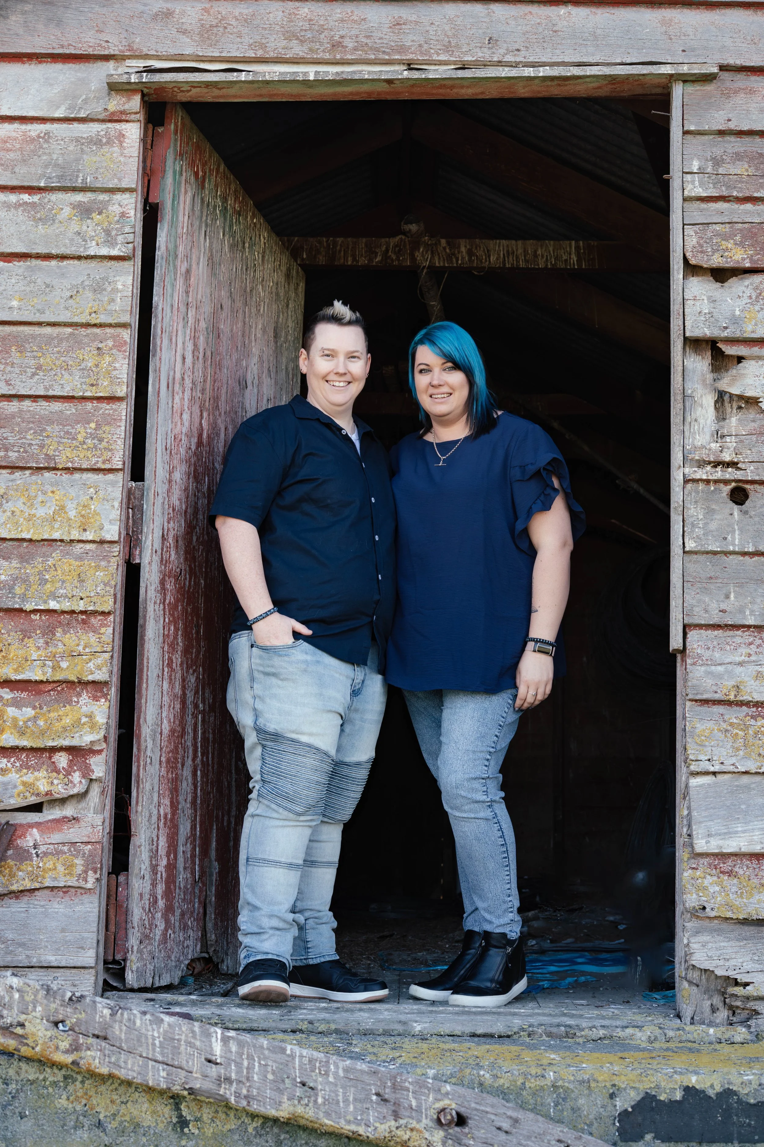 Two women with short and blue hair, wearing dark blue shirts and jeans, smiling and standing inside a weathered, old barn.'