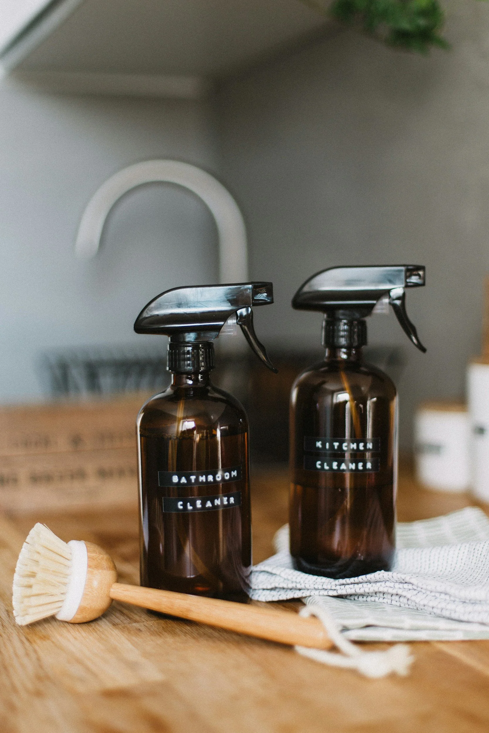 Two brown glass bottles labeled 'Bathroom Cleaner' and 'Kitchen Cleaner' with black spray nozzles, placed on a wooden surface with a textured white cloth, a wooden brush, and a cardboard box in the background.