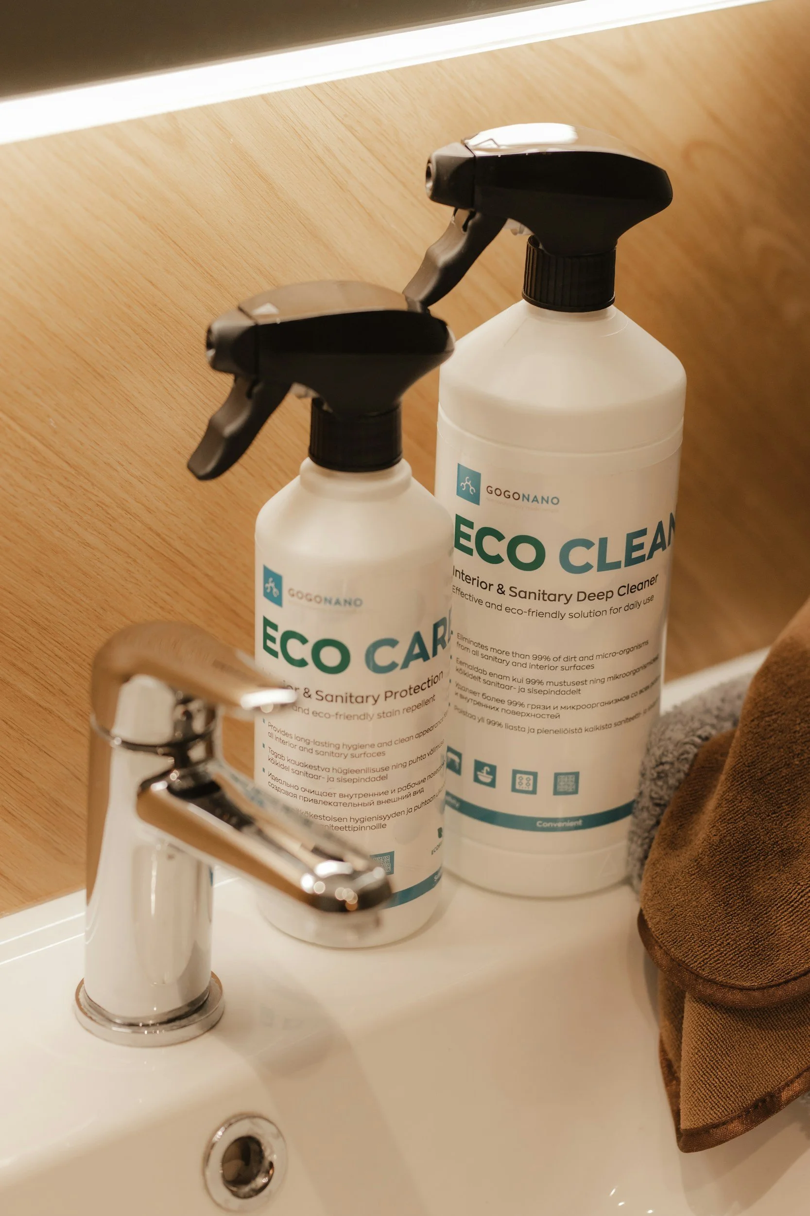 Two bottles of ECO CLEAN interior and sanitary deep cleaner placed on a bathroom sink countertop next to a silver faucet and a brown towel, with a wooden wall in the background.