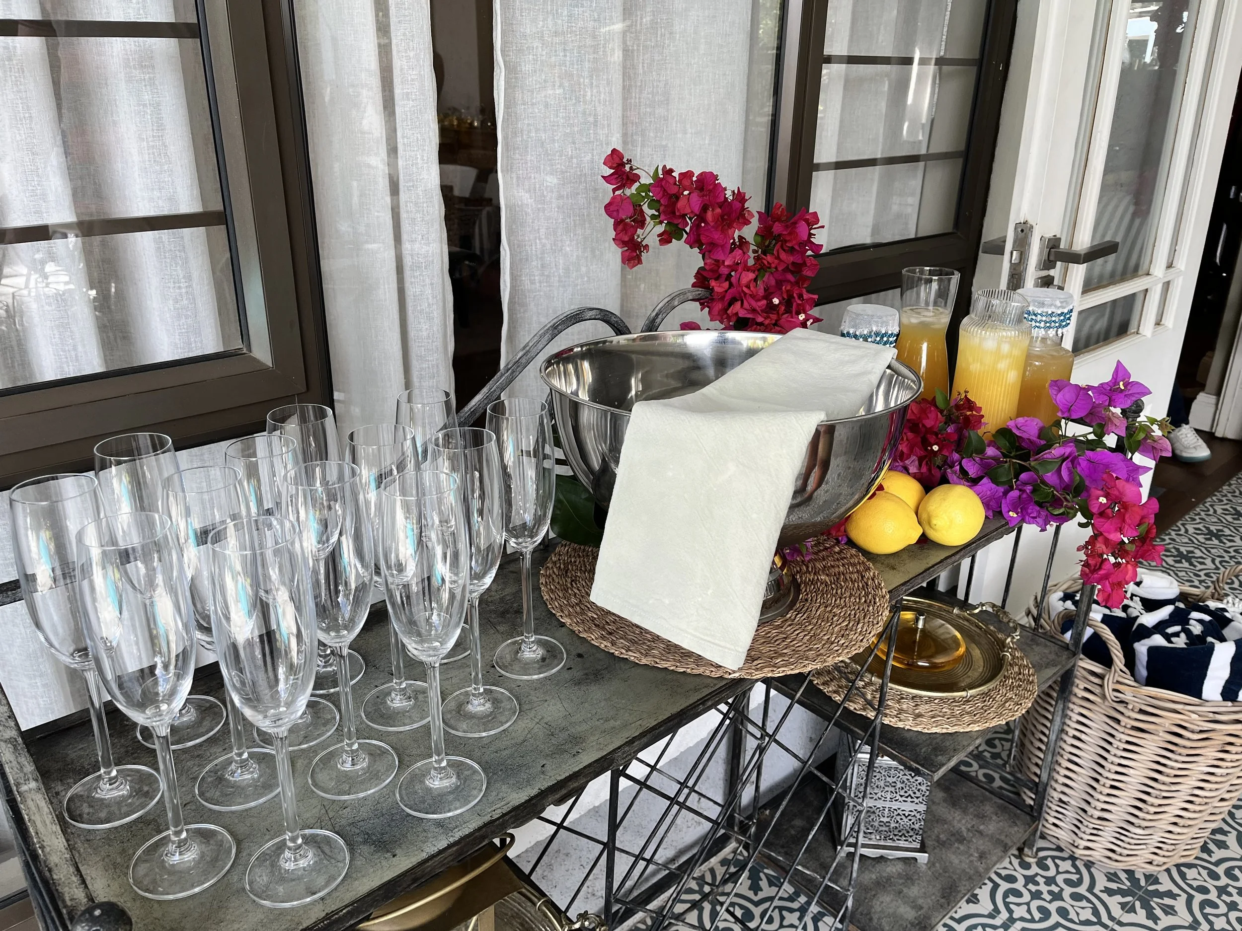 A table set with multiple empty champagne flutes, a silver punch bowl with a white towel, and a tray of yellow lemons surrounded by pink bougainvillea flowers and several pitchers of orange juice. The background shows a window with white curtains and