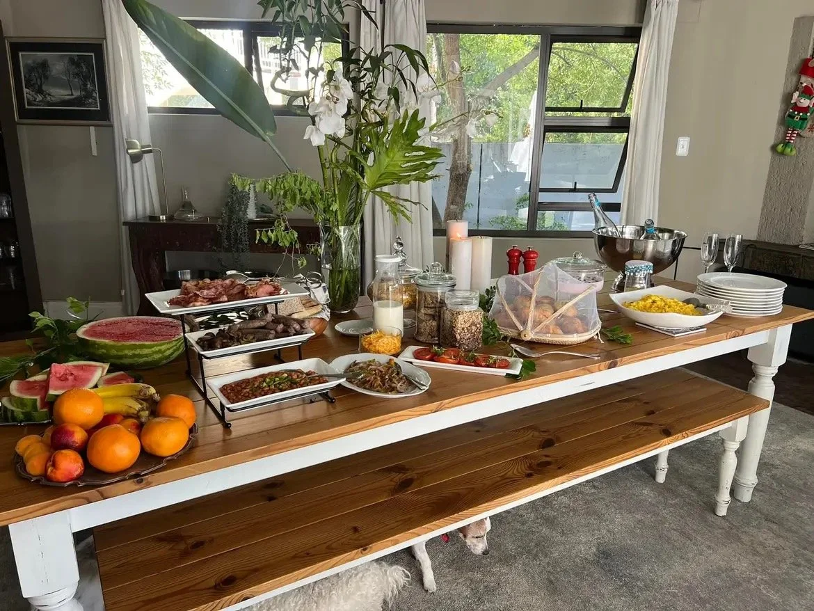 A wooden dining table set with various dishes, fruits, and beverages for a meal, with a large window and house plants in the background.