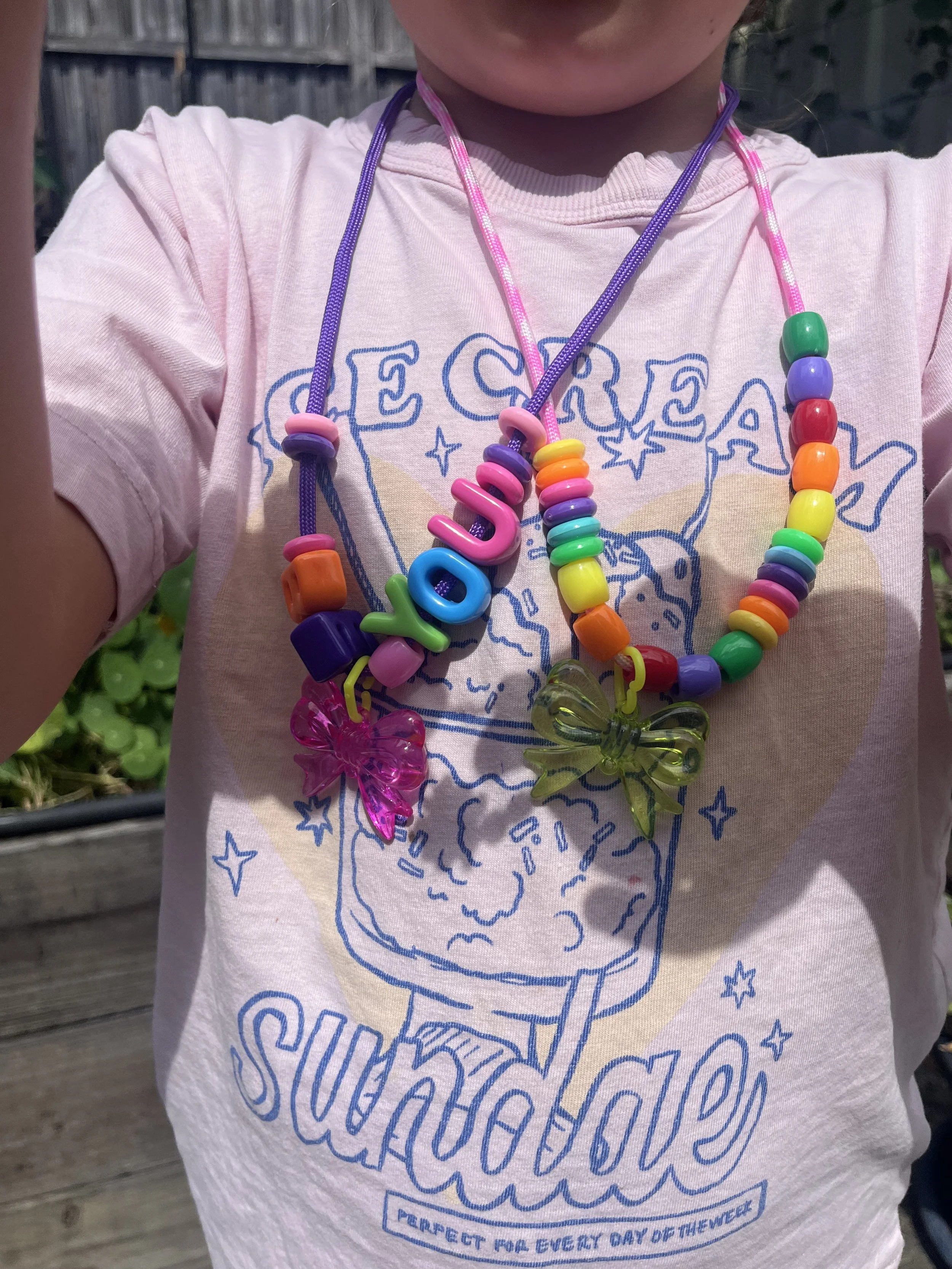 Close-up of a child's chest wearing a pink T-shirt with a blue graphic and text, and rainbow-colored beaded necklaces with butterfly-shaped charms. The child is outdoors with a wooden fence and greenery in the background.