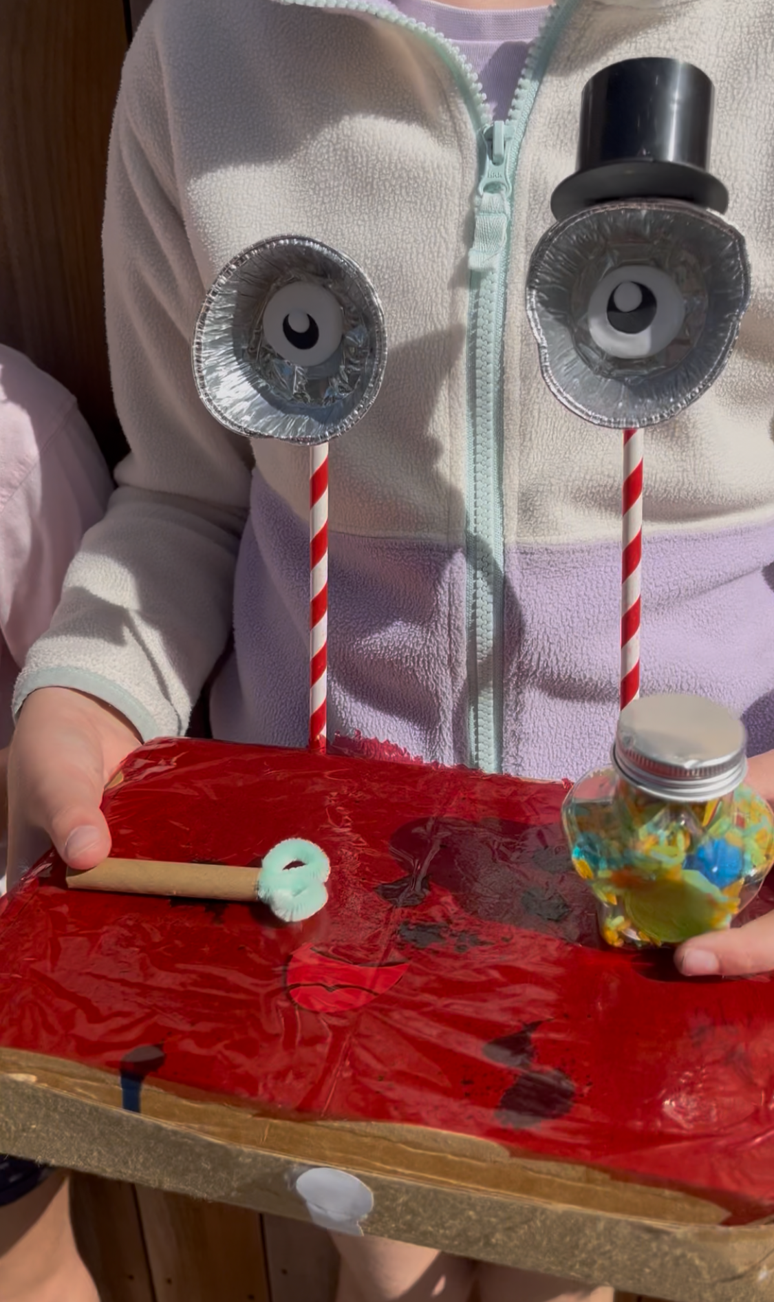 Person holding a craft project with two handmade owl figures made from black cups, paper eyes, and striped straws on a red table.