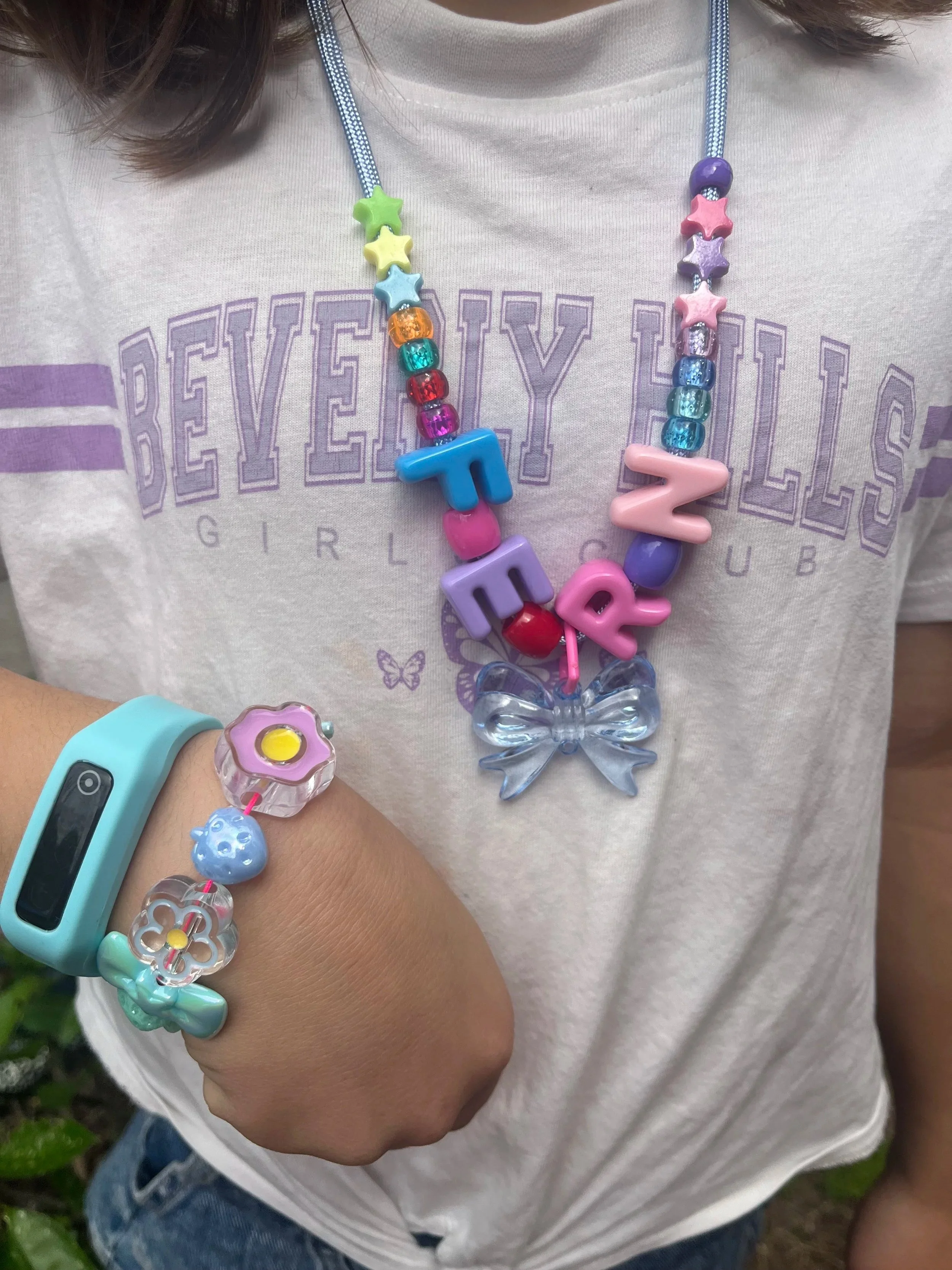 Close-up of a young girl wearing colorful jewelry, including a beaded necklace with stars and the word 'LOVE', a bracelet with flower charms, and a wearable device on her wrist. She is wearing a light-colored T-shirt with the word 'REVEALY' partly vi