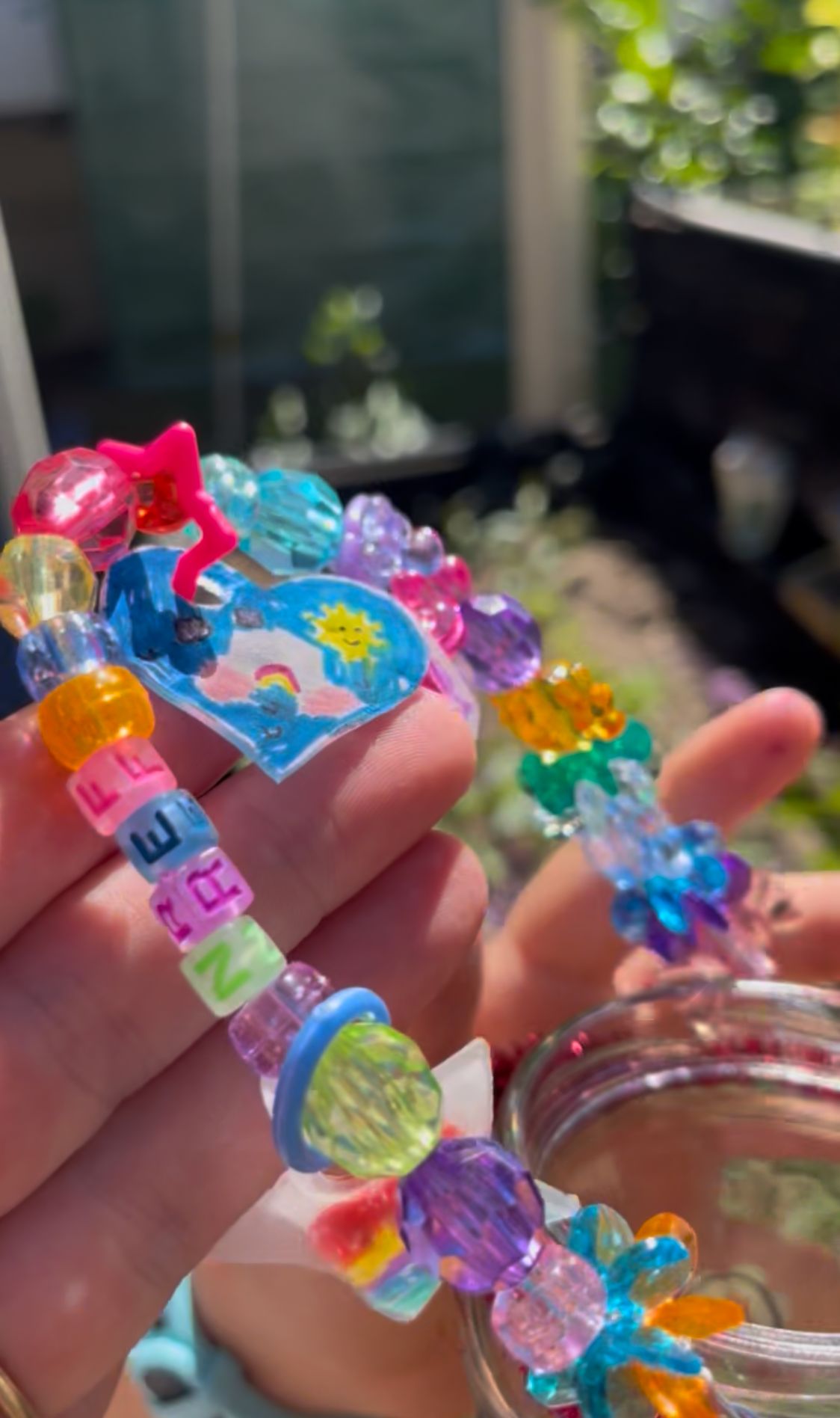 Close-up of a colorful beaded bracelet with letter beads, a small heart sticker with a rainbow, sun, and clouds, and a pink star-shaped bead, held in a person's hand outdoors with a blurred background of greenery and a building.