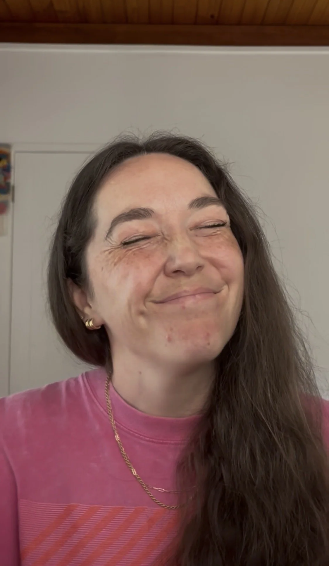 A woman with long brown hair, closed eyes, and a big smile, wearing a pink shirt and gold earrings. She appears to be happy or laughing.