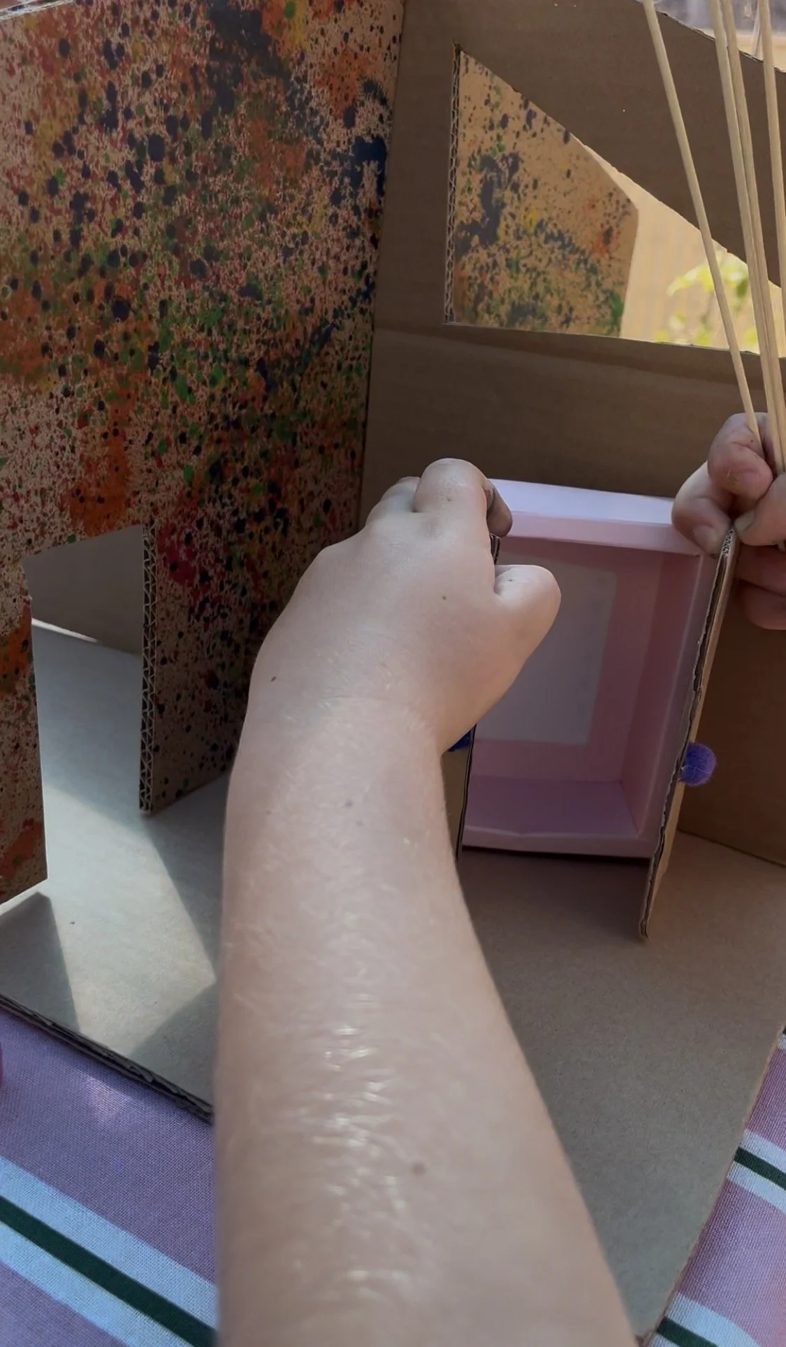 Person assembling a cardboard box with a pink interior, with colorful patterned cardboard pieces around, on a striped tablecloth.