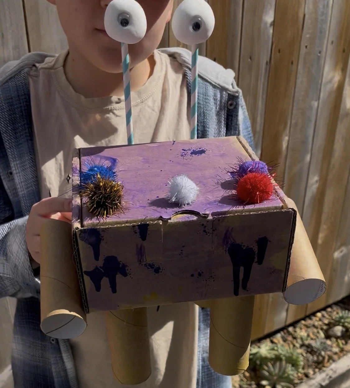 Child holding a homemade robot craft made from a cardboard box with googly eyes on straws, pipe cleaners, and fur pompoms.