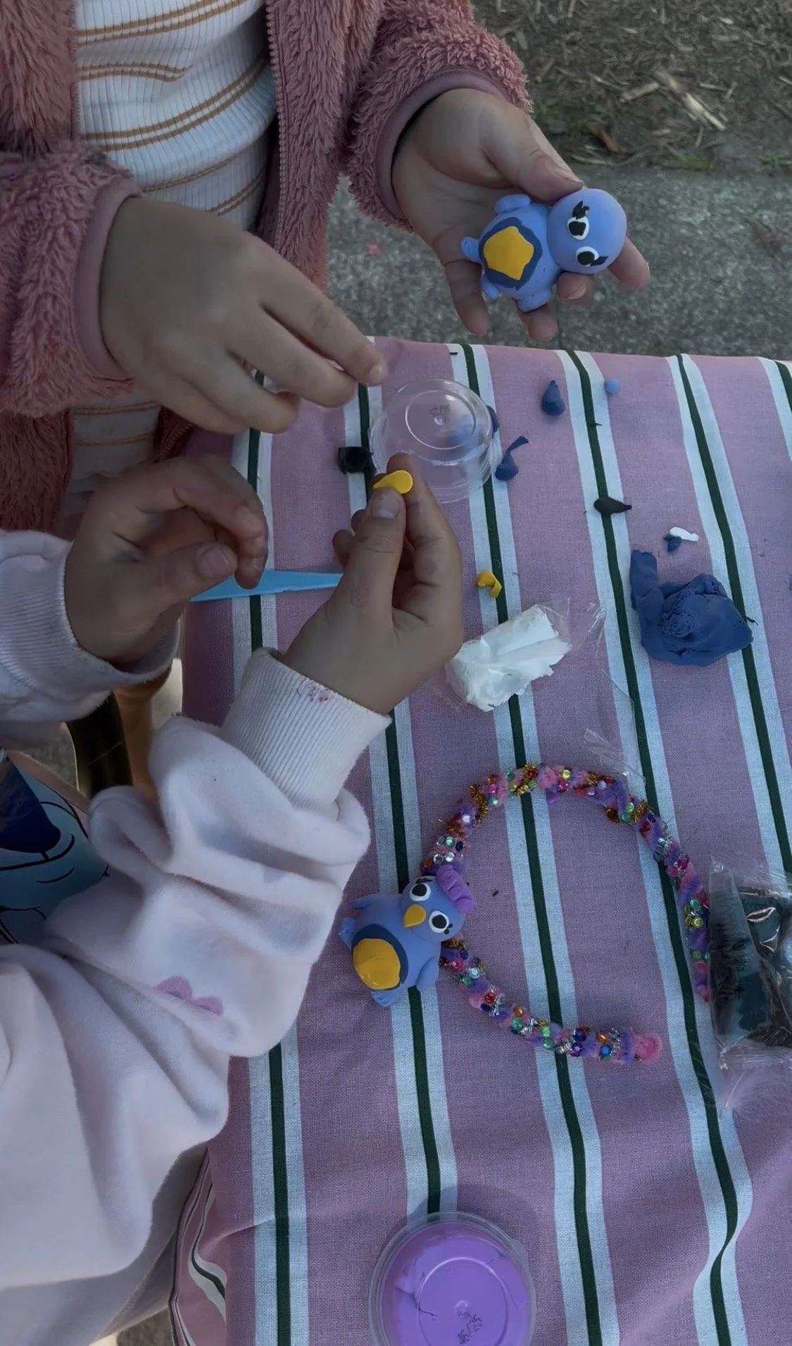 Children creating colorful slime and decorating toy figures at a table with a pink, green, and white striped tablecloth.