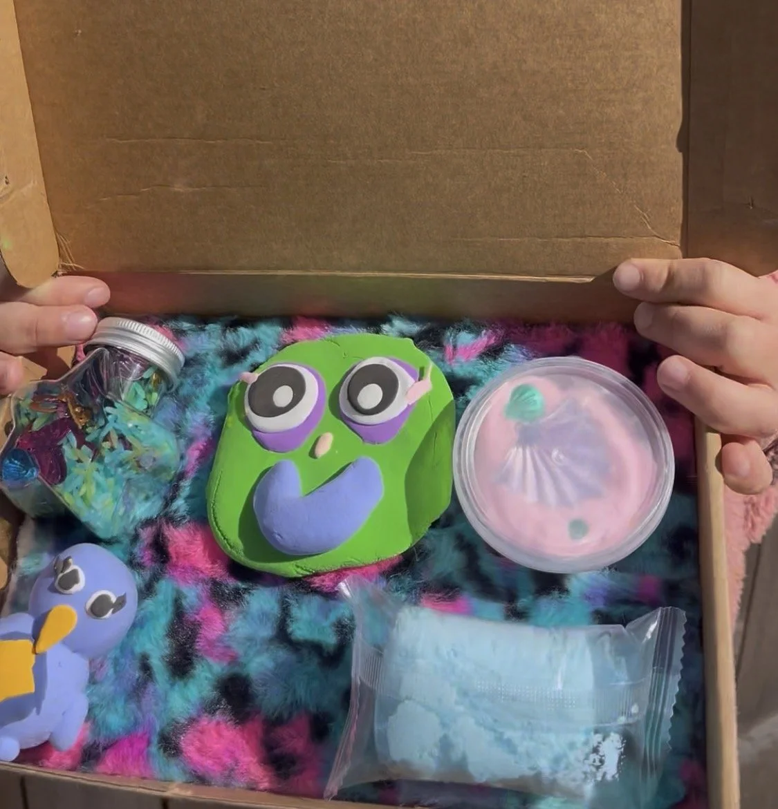 A box filled with colorful craft supplies, including a green and purple painted rock with large eyes, a small blue and yellow painted rock, a container of pink slime, a jar with small pieces of colored paper, and a plastic bag of blue craft powder, a