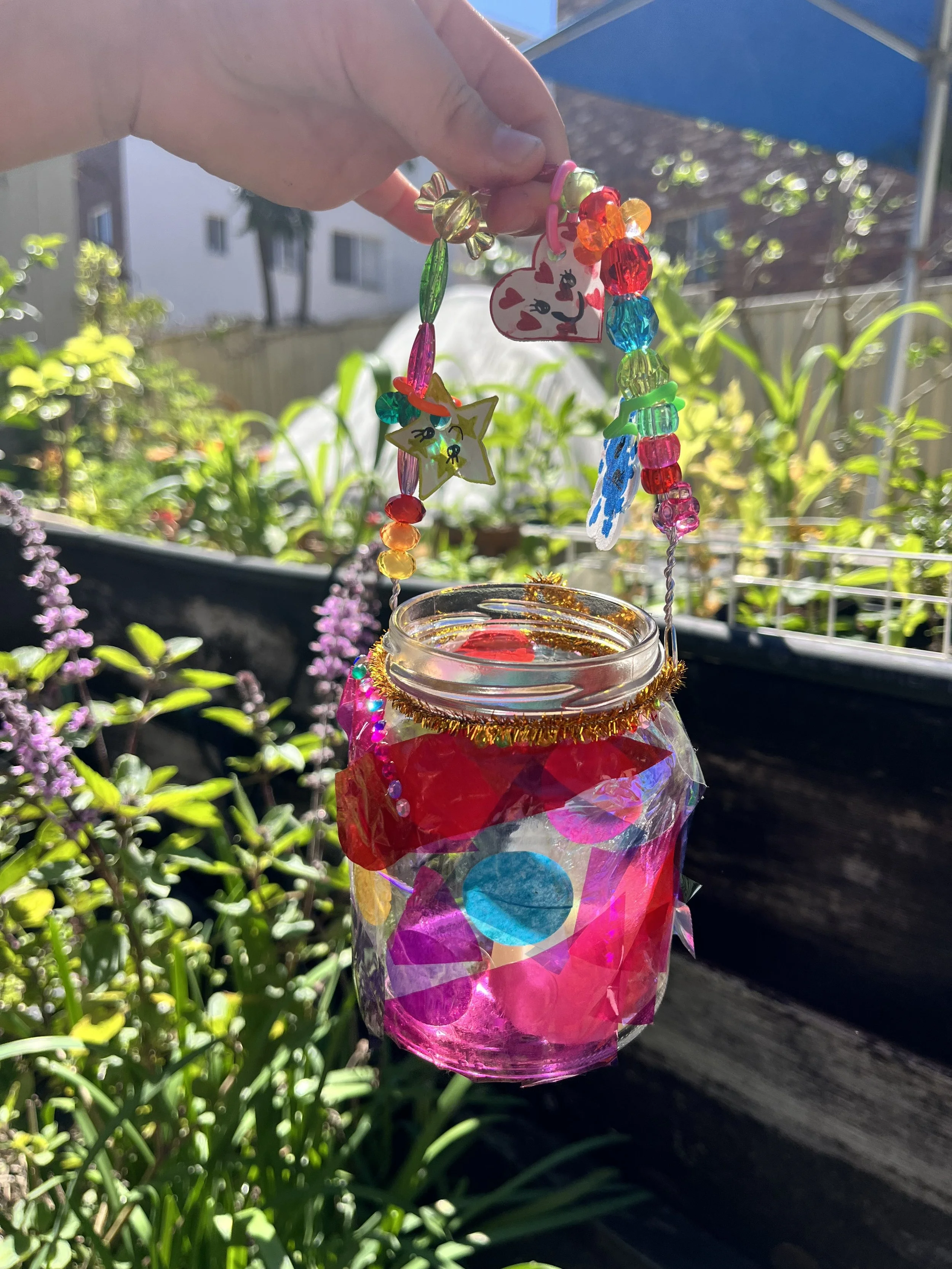 Colorful beaded decoration hanging from a glass jar with bright sunlight, a garden with green plants and purple flowers in the background.