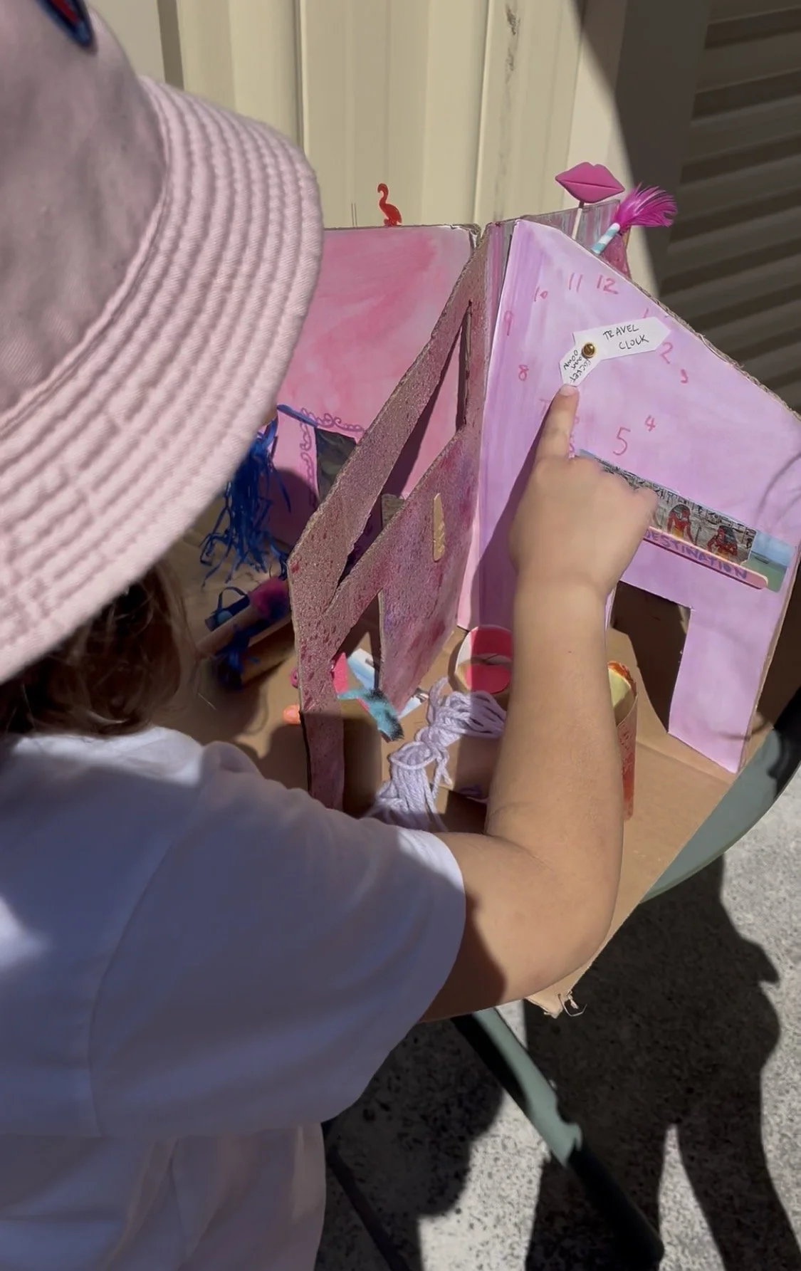 A young girl wearing a pink hat and white shirt is looking at a pink, handmade, cardboard dollhouse with a pink clock and various decorations inside.