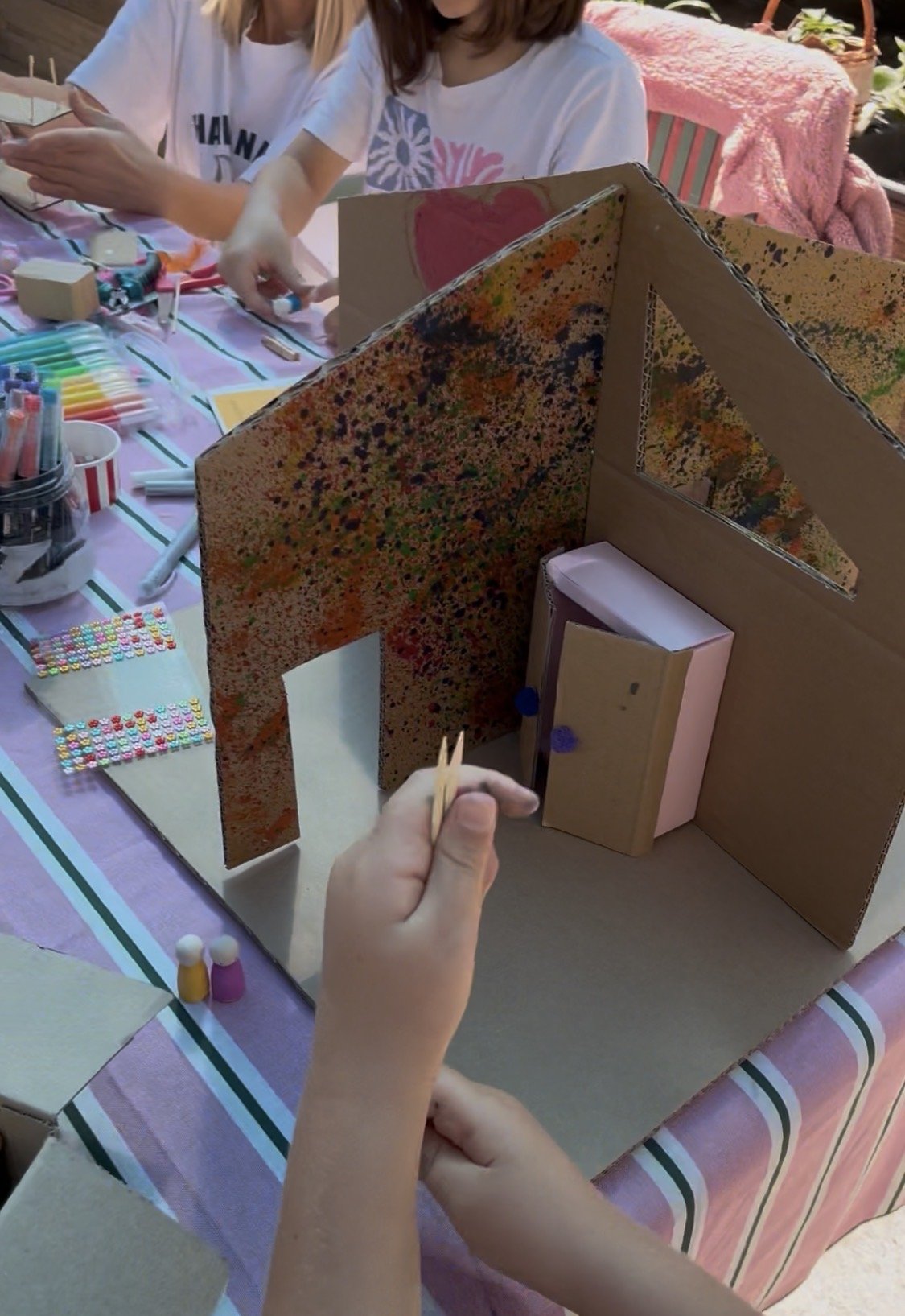 Children creating a miniature house model with cardboard, paint, and small decorations on a table covered with a pink-striped tablecloth.