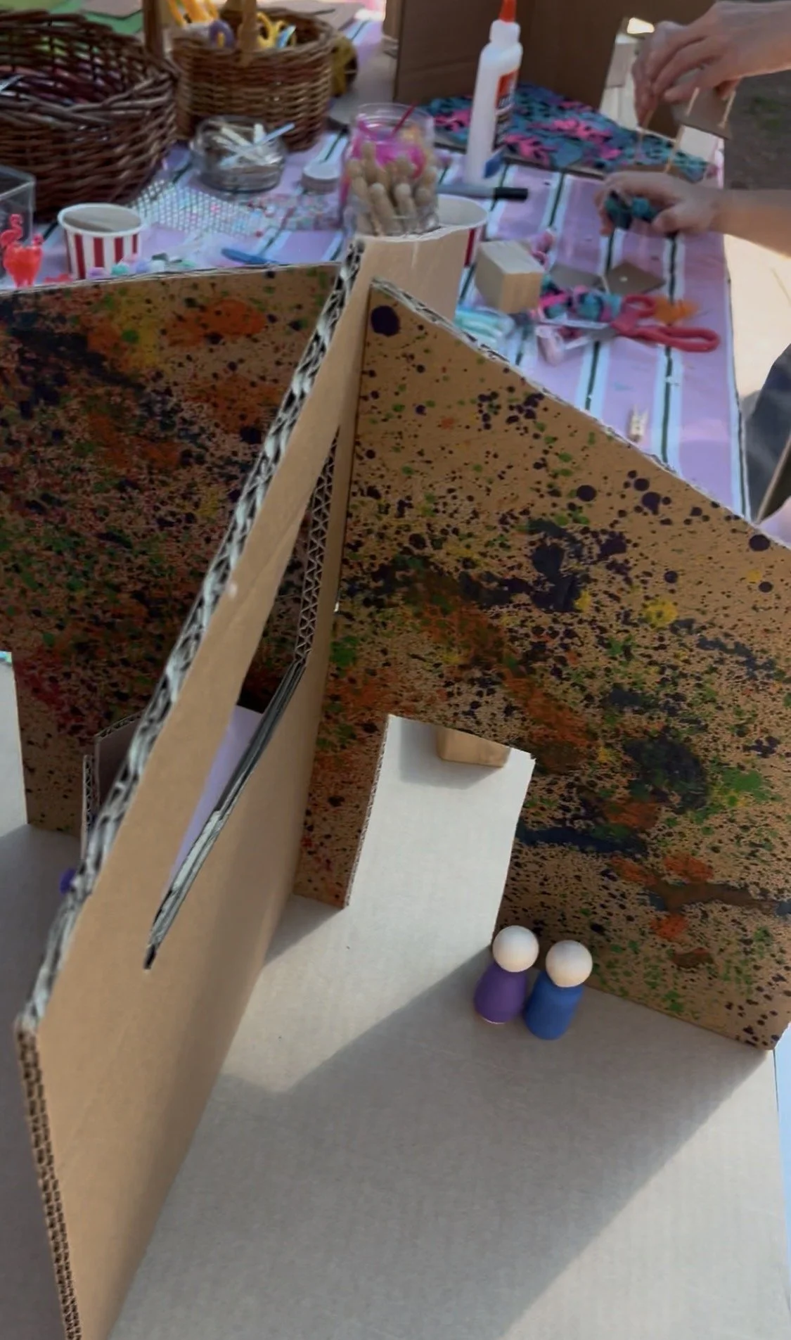A craft project setup with a cardboard structure, paint splattered on it, and craft supplies including scissors, glue, and small figurines. The scene appears to be a workspace for assembling a miniature model or diorama.