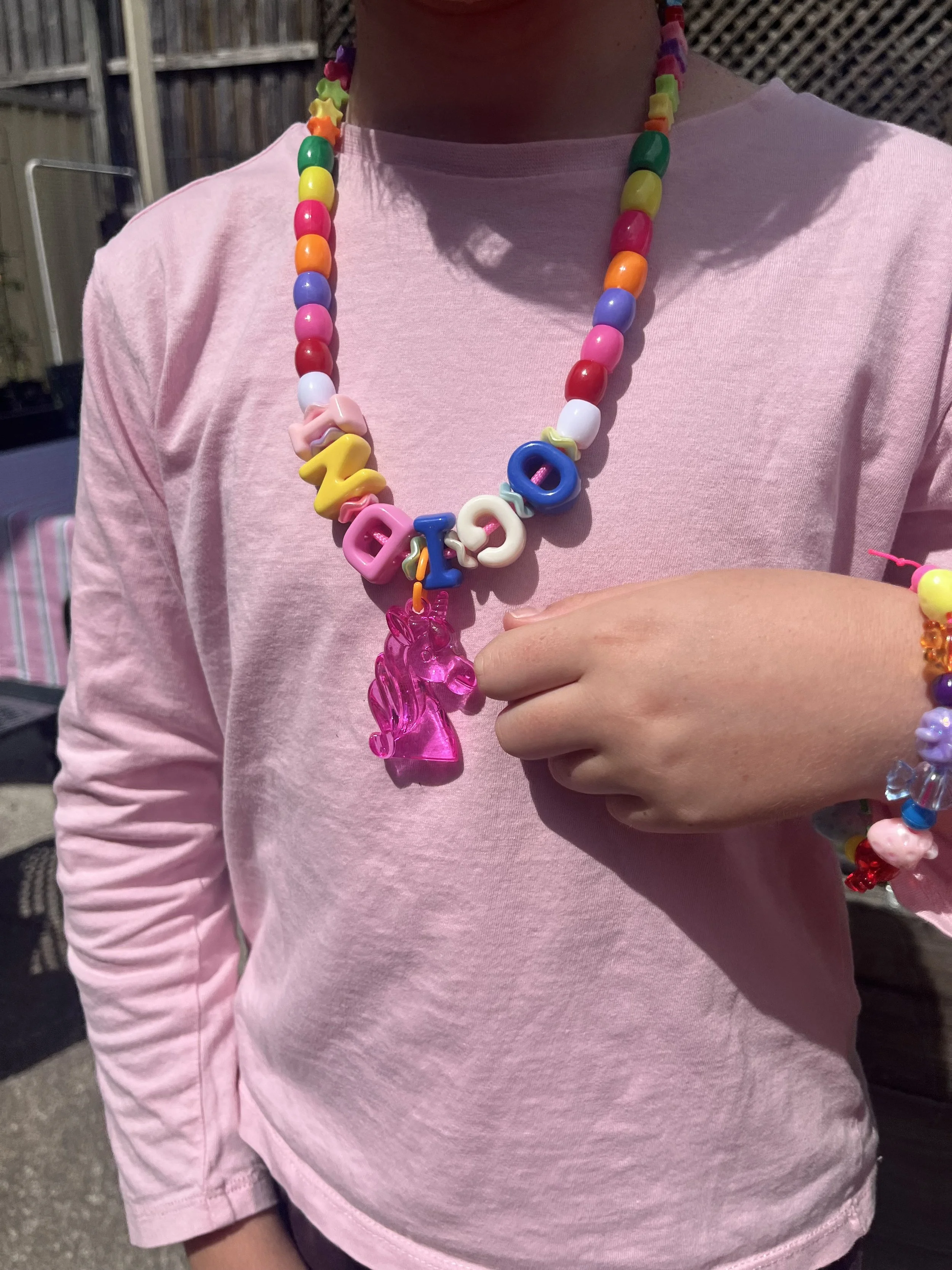 Person wearing a pink shirt and colorful beaded necklace with alphabet beads spelling 'LOVE'. They are holding a pink plastic figure. The background includes a wooden fence and outdoor furniture.
